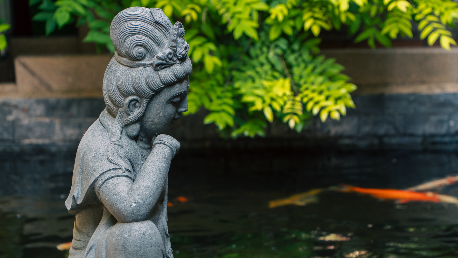 A tranquil stone statue by a pond with colorful koi fish and lush greenery.