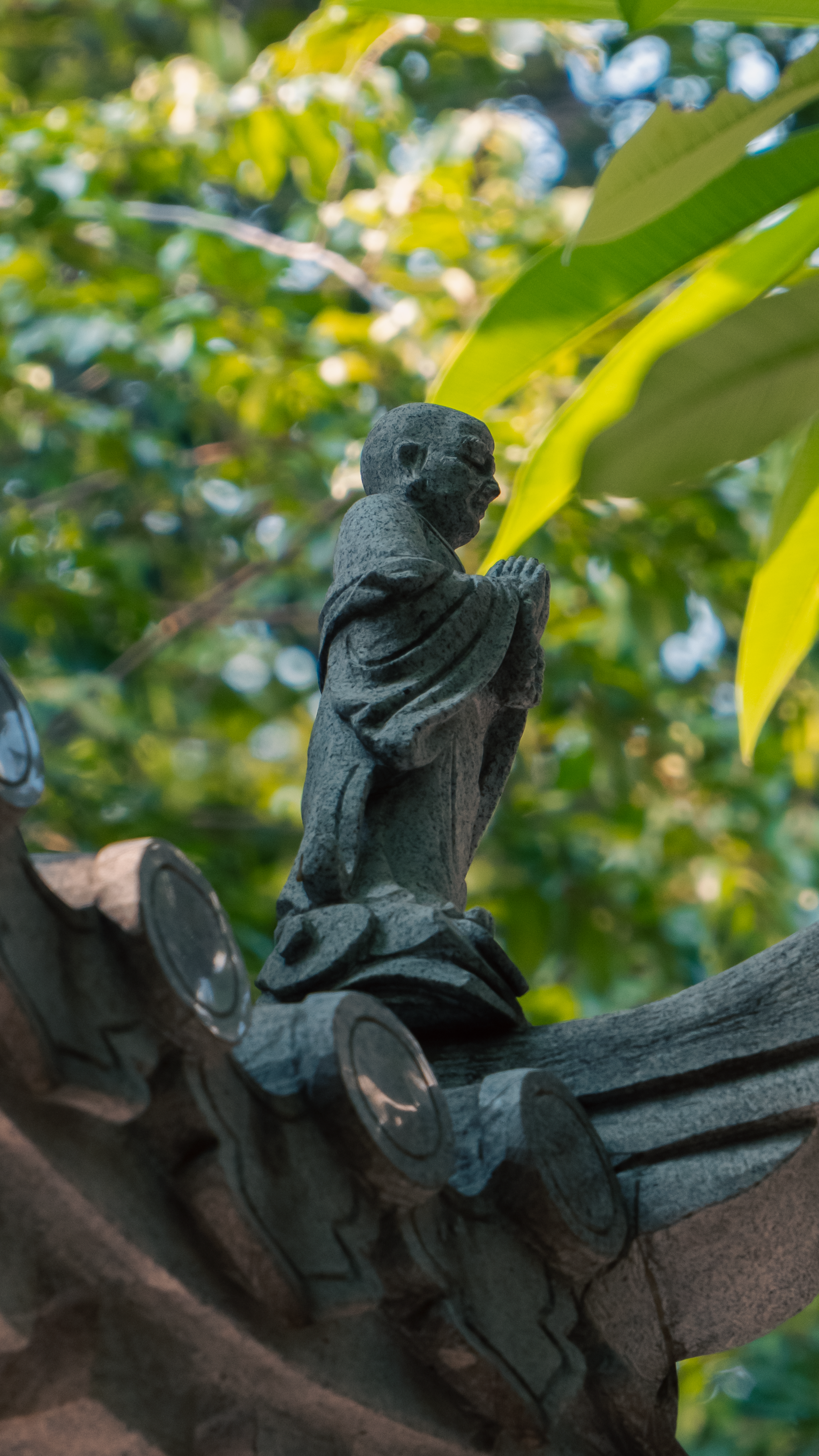 A stone figure adorns a roof, surrounded by lush green foliage, creating a serene atmosphere.