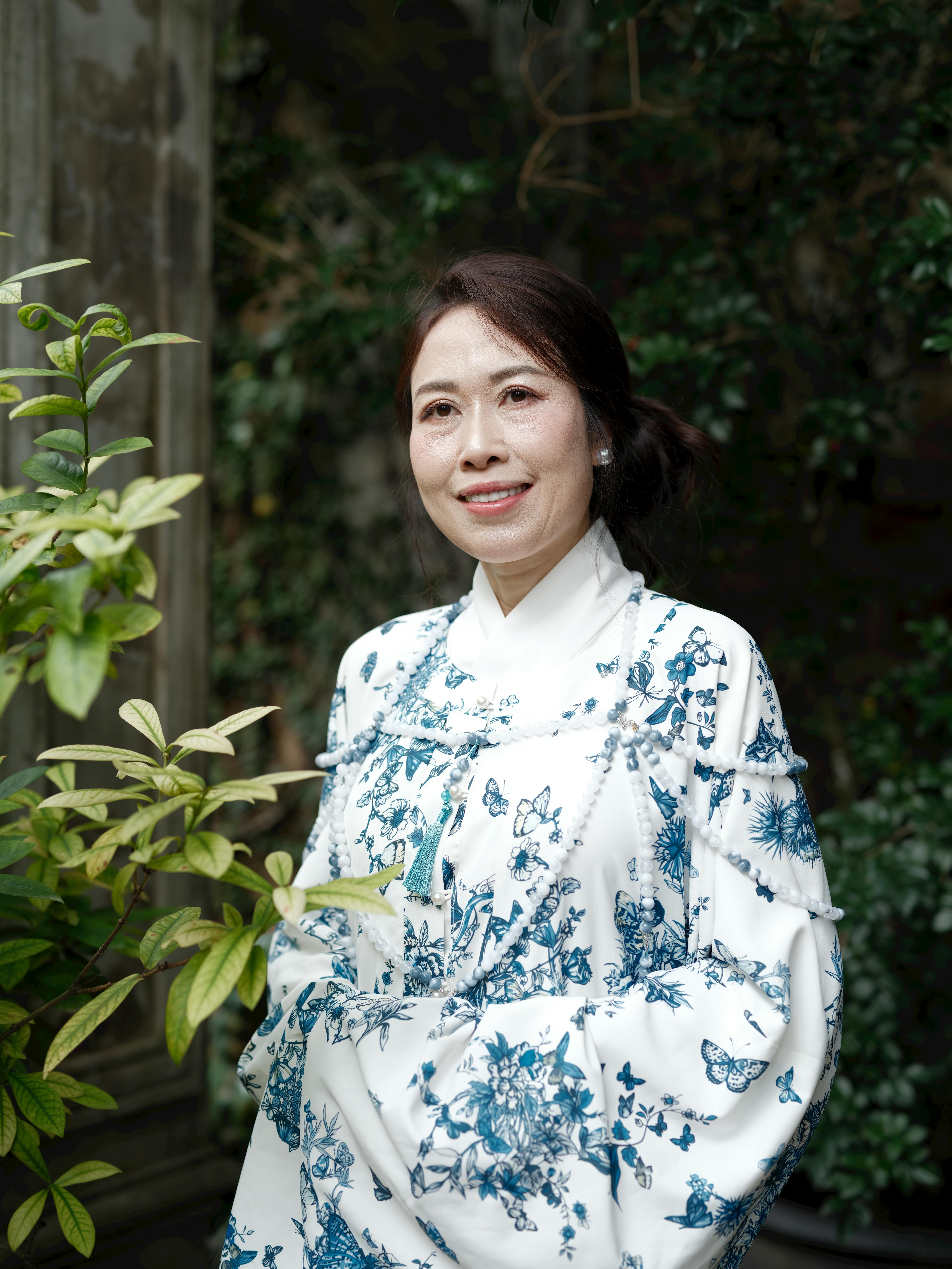 Smiling woman in traditional floral attire standing outdoors surrounded by lush greenery.