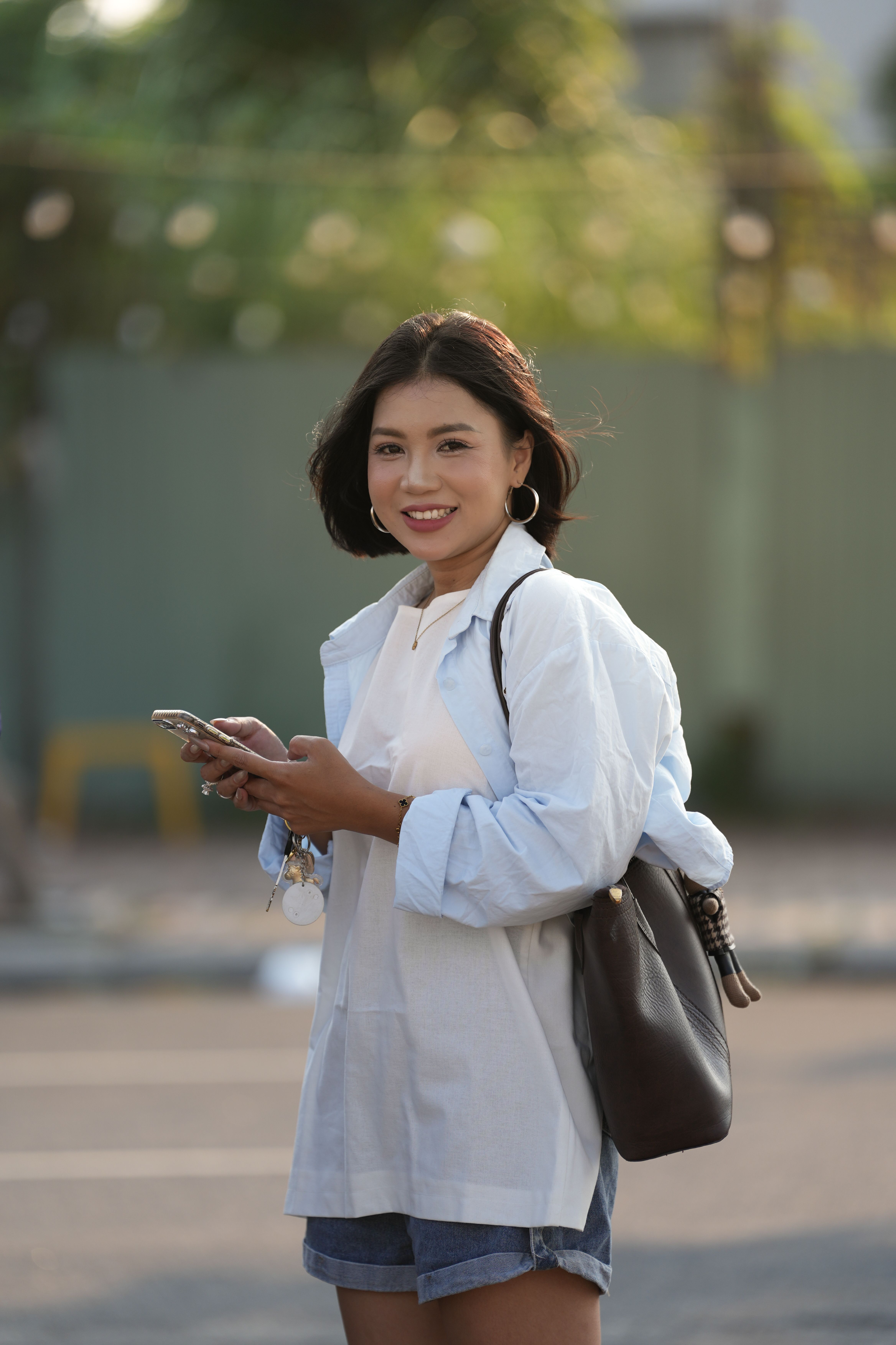 Smiling woman in casual attire with smartphone on a sunny day outdoors.