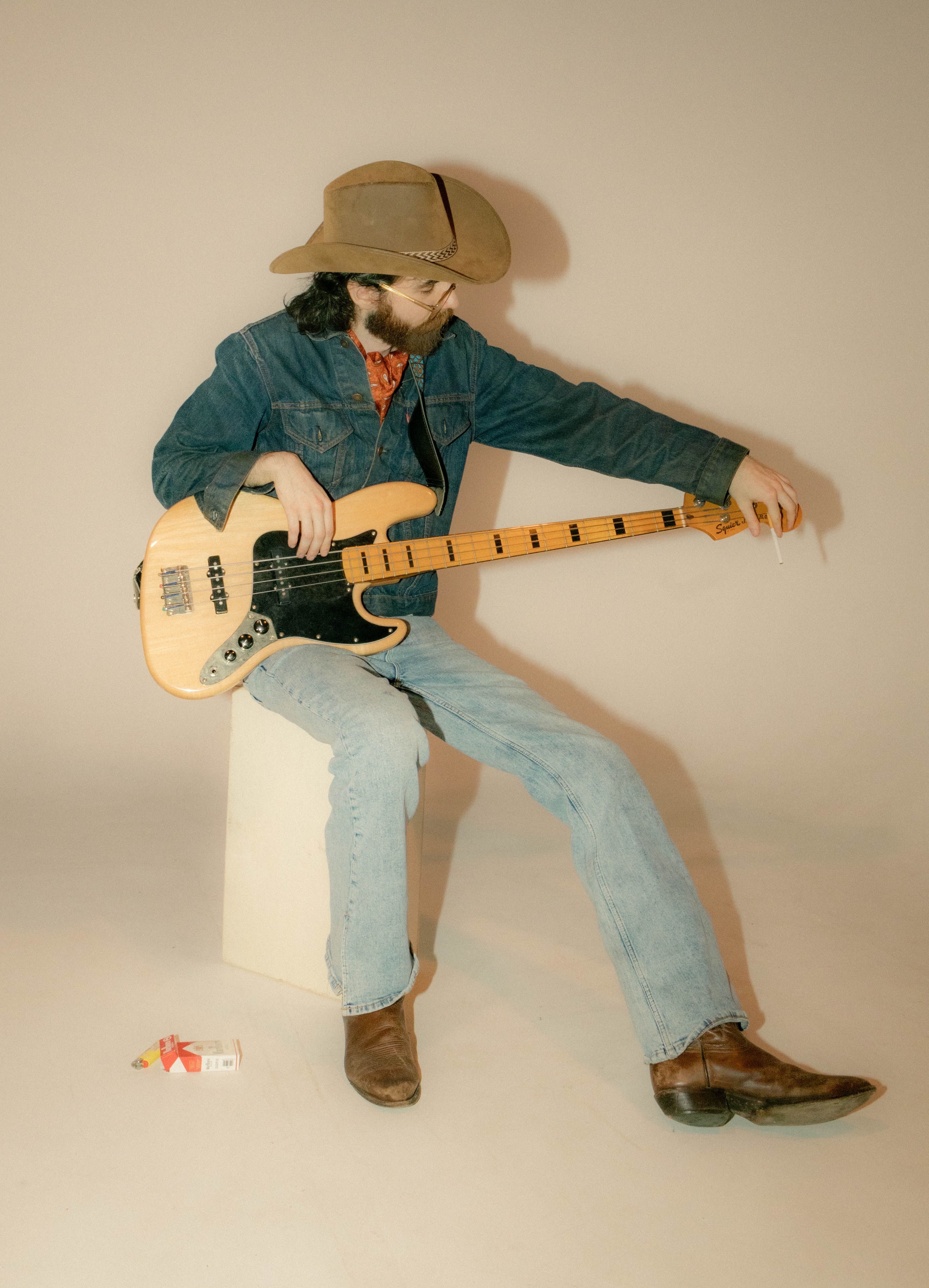 A cowboy musician tuning a vintage electric guitar, sitting on a stool in a studio setting.