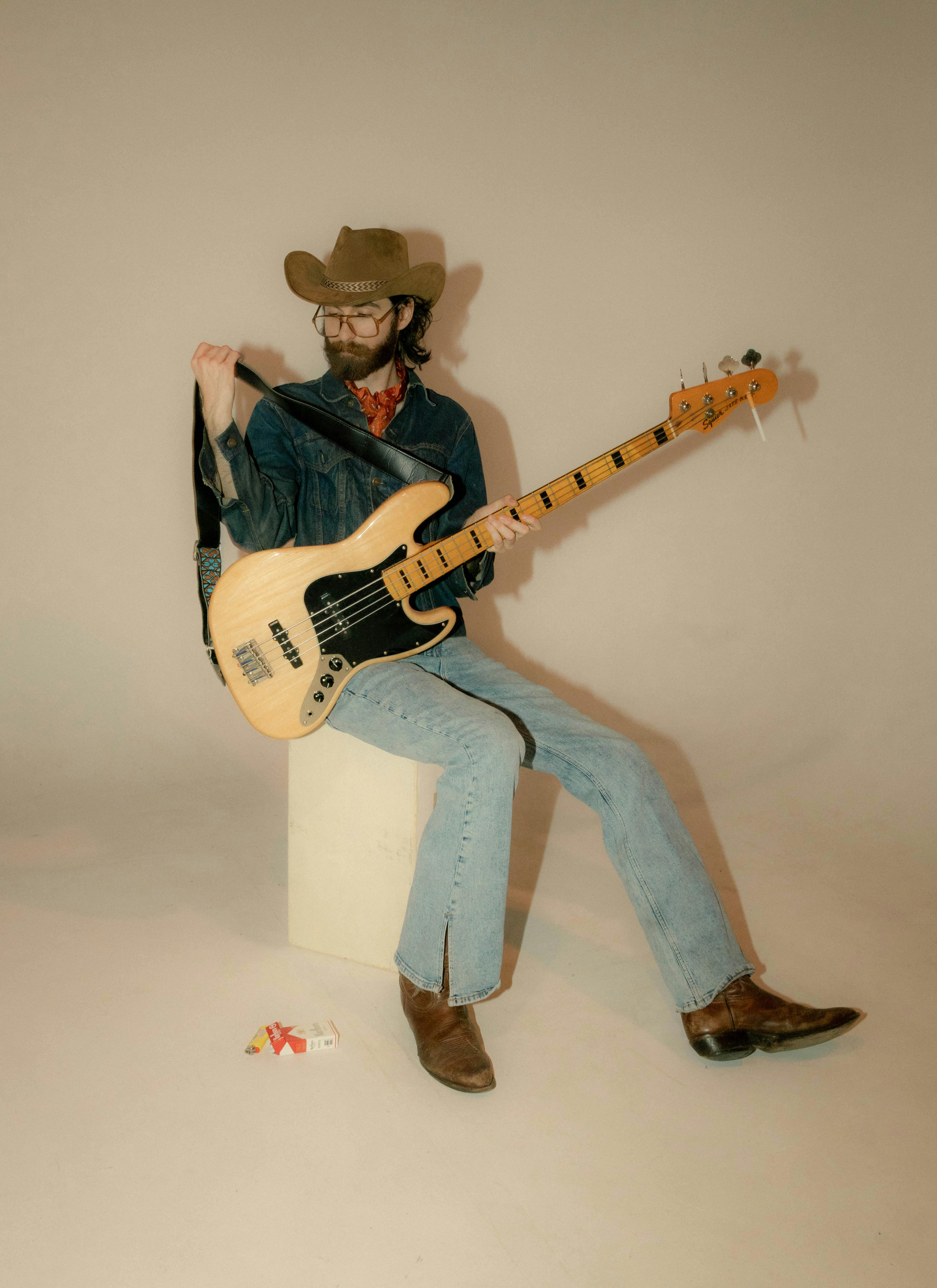Man in cowboy hat and denim playing electric bass guitar indoors.
