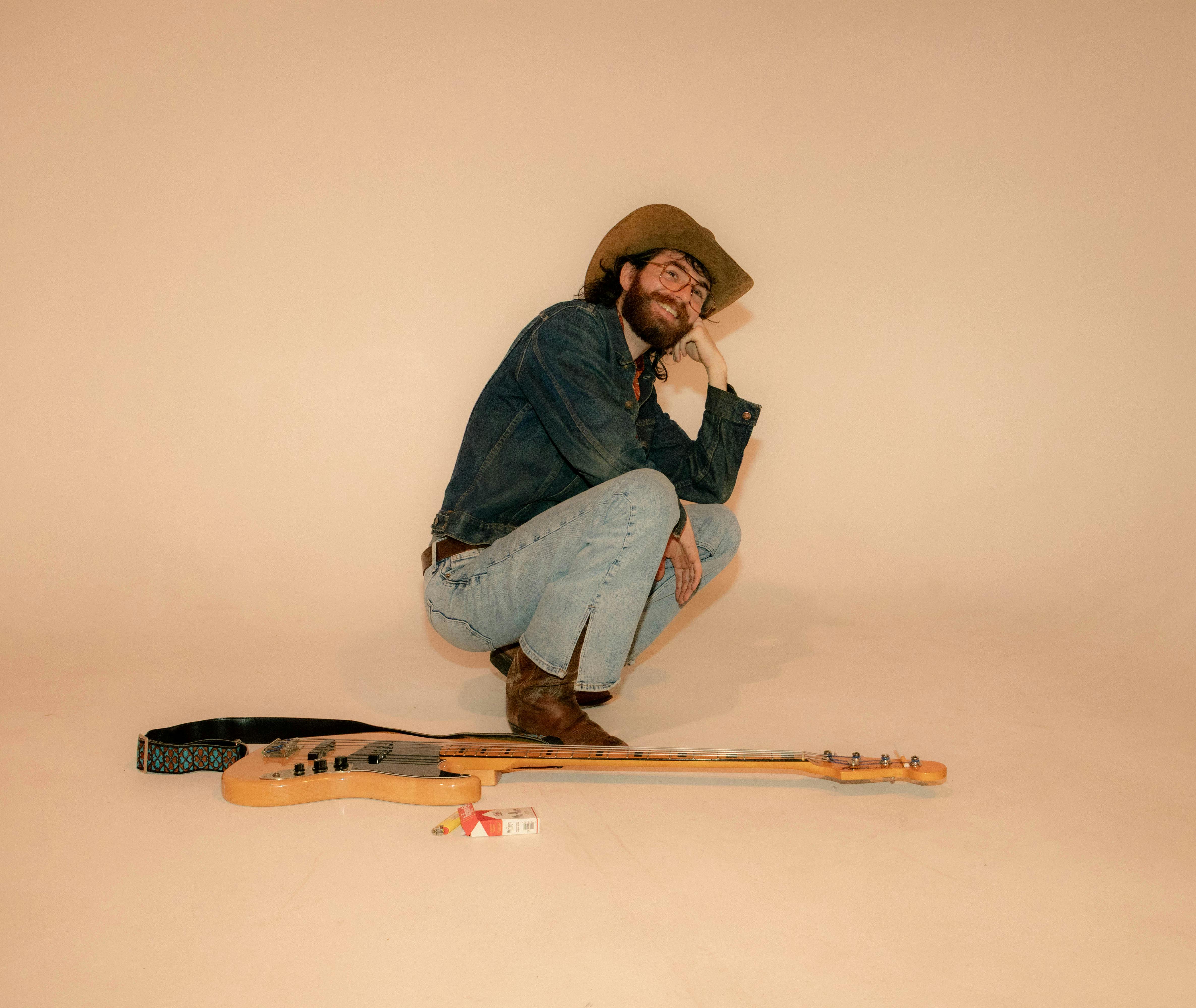 A stylish man in denim and cowboy hat crouches beside an electric guitar in a neutral-toned studio.