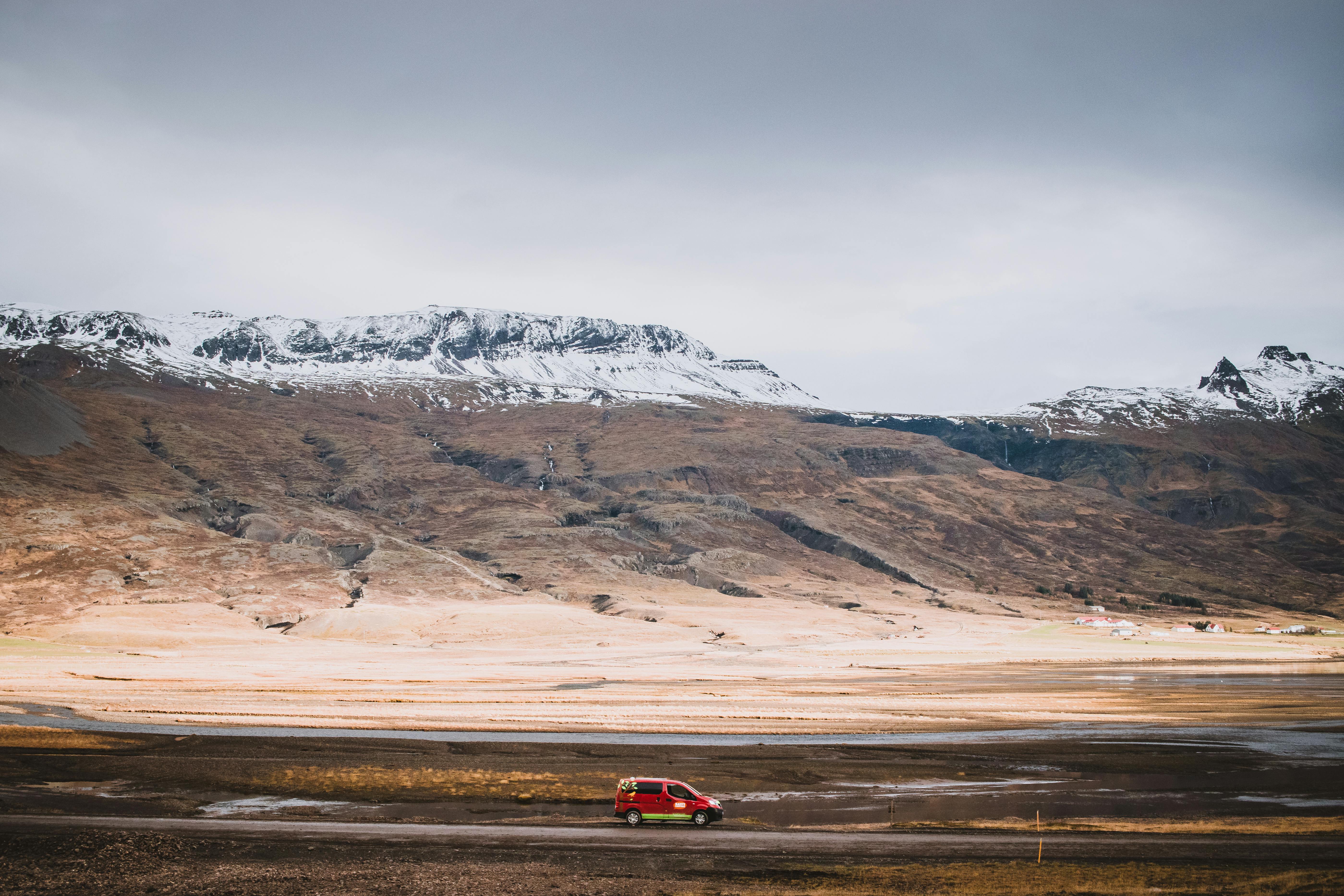 A red car travels through Iceland's rugged landscape with snowy mountains under a cloudy sky.