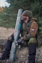 一名身着迷彩服的士兵手持大型武器，坐在森林中准备战斗