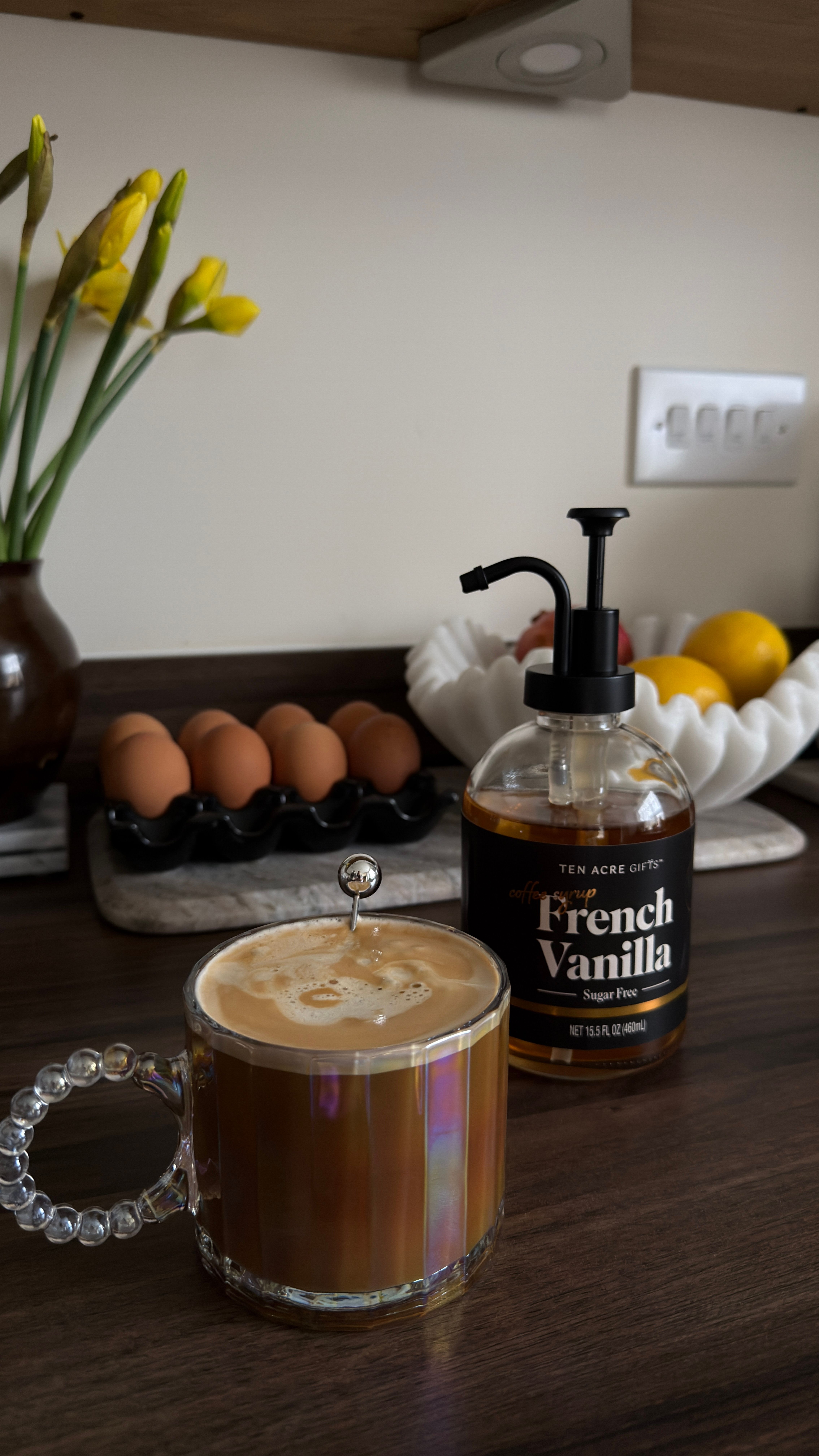 A glass bottle of golden vanilla syrup being poured into a latte, with a scraped vanilla bean pod and a small dish of sugar sitting next to it.