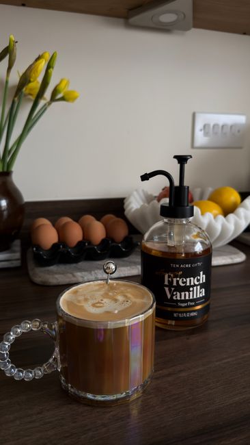 A glass bottle of golden vanilla syrup being poured into a latte, with a scraped vanilla bean pod and a small dish of sugar sitting next to it.