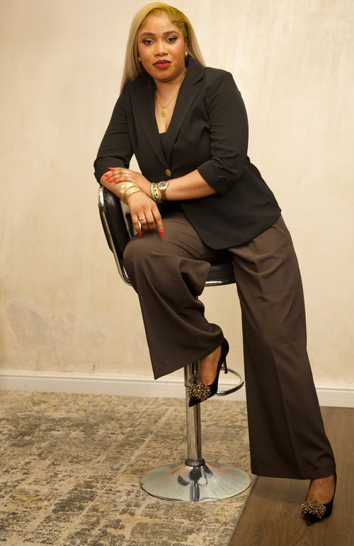 Free A confident woman in a black blazer and brown trousers seated on a stool indoors, exuding professionalism. Stock Photo