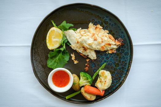 A beautifully plated meal featuring almond-crusted fish, vegetables, and sauce on a dark plate.