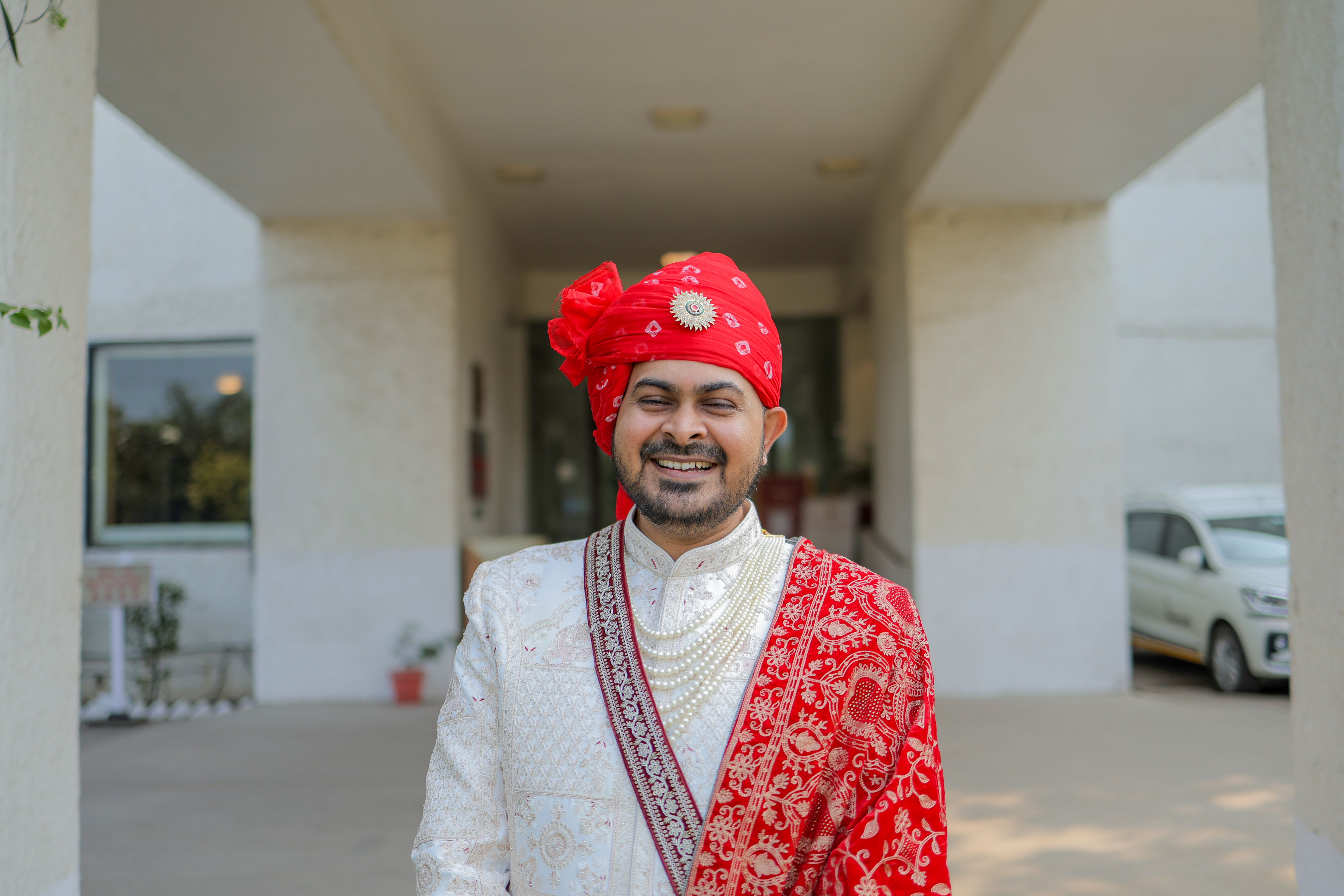 A smiling groom in intricate red and white traditional Indian attire outdoors, showcasing rich cultural fashion.