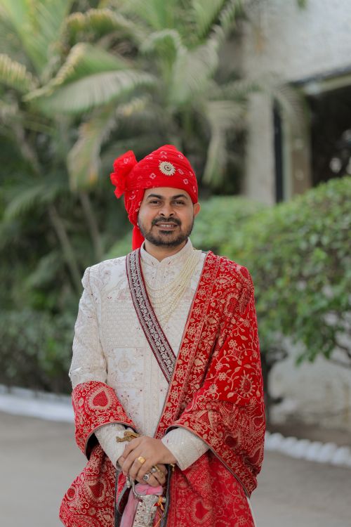 Free Indian groom wearing traditional attire with red turban and embroidered shawl outdoors. Stock Photo