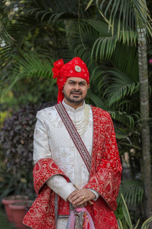 Free Indian groom dressed in red and white traditional attire with ornate jewelry standing outdoors. Stock Photo