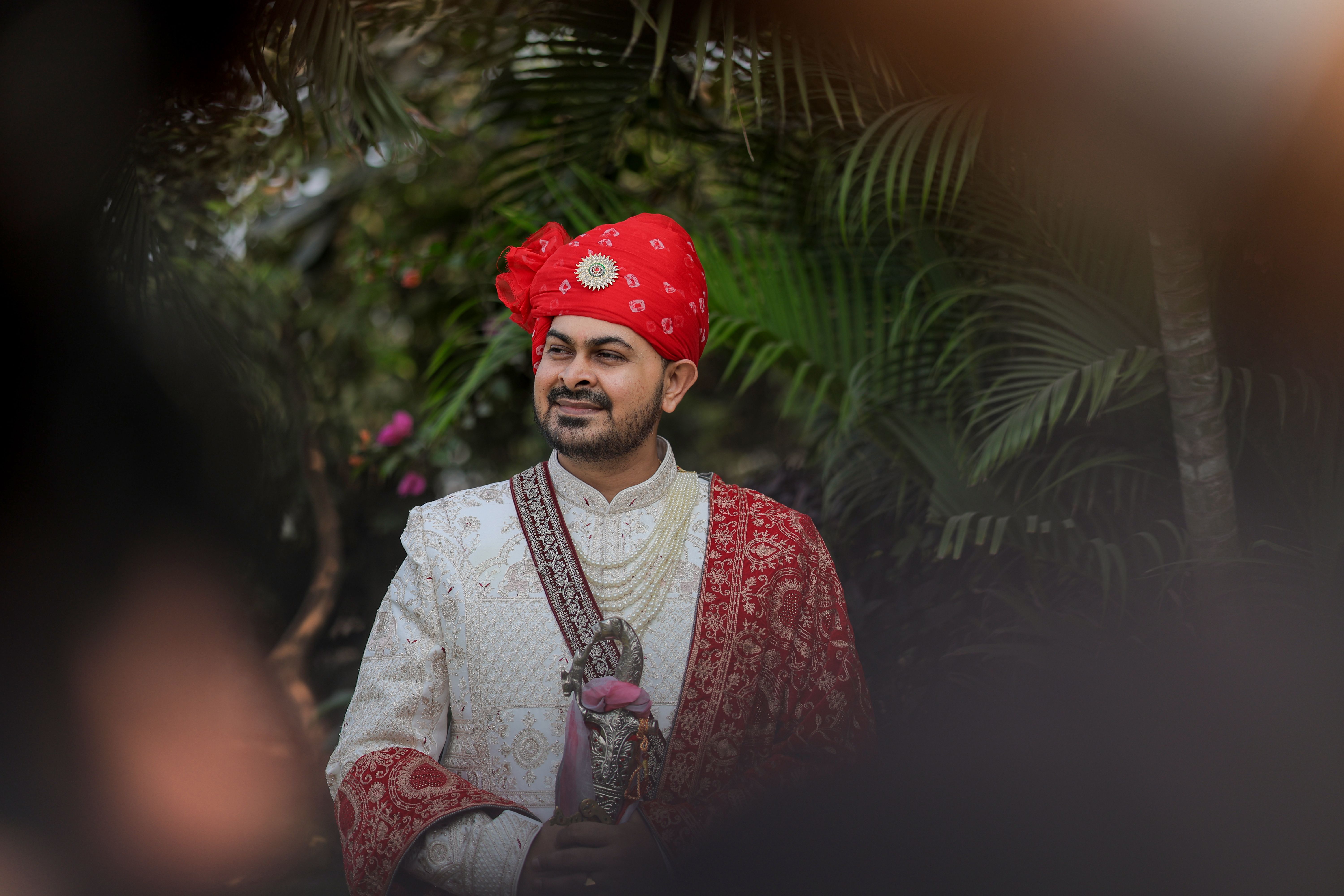 A South Asian groom in traditional attire with red turban and ornate white sherwani, posing outdoors.