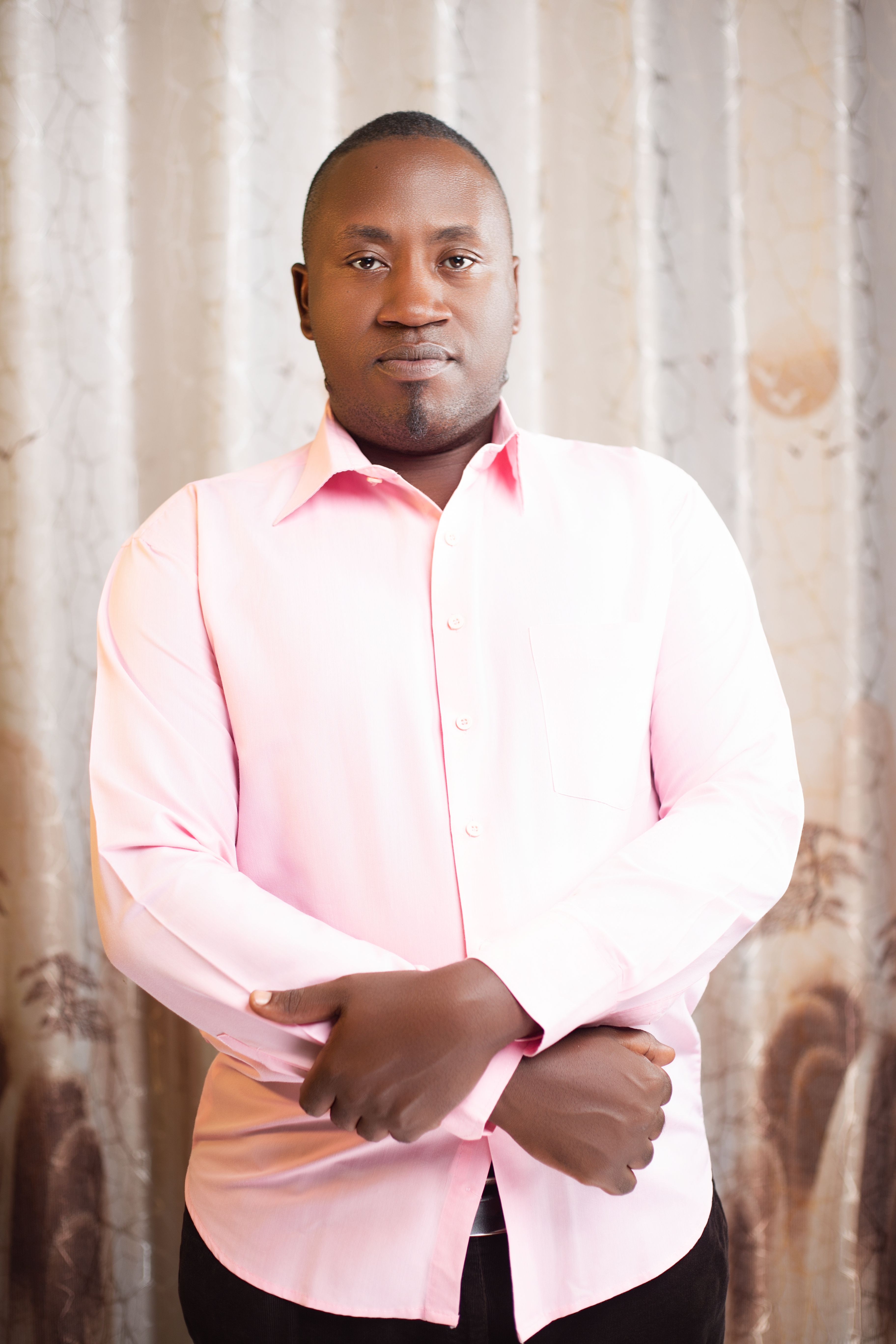 A confident man stands with folded arms indoors, wearing a stylish pink shirt.