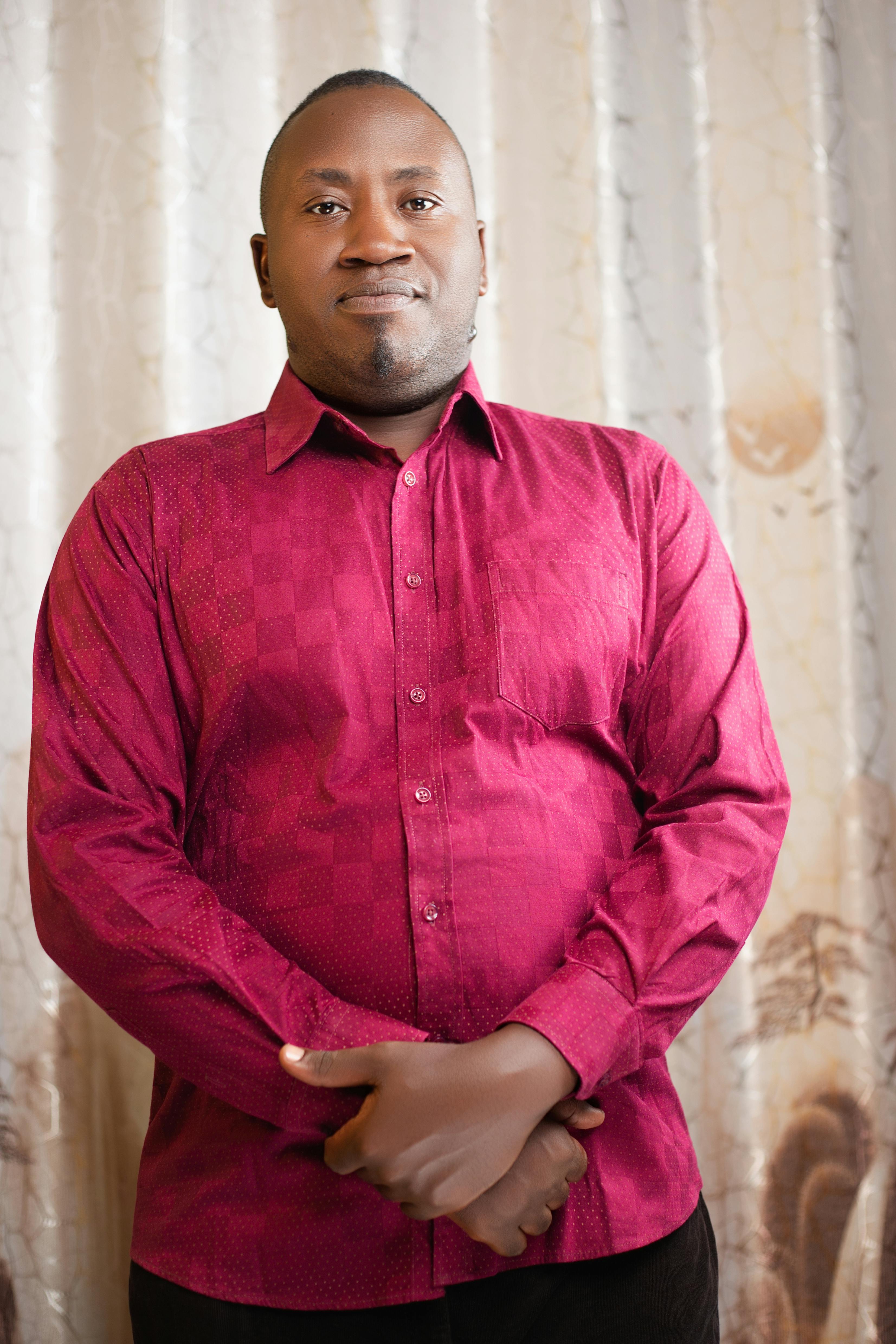 A confident man poses indoors wearing a vibrant red shirt with a subtle patterned backdrop.