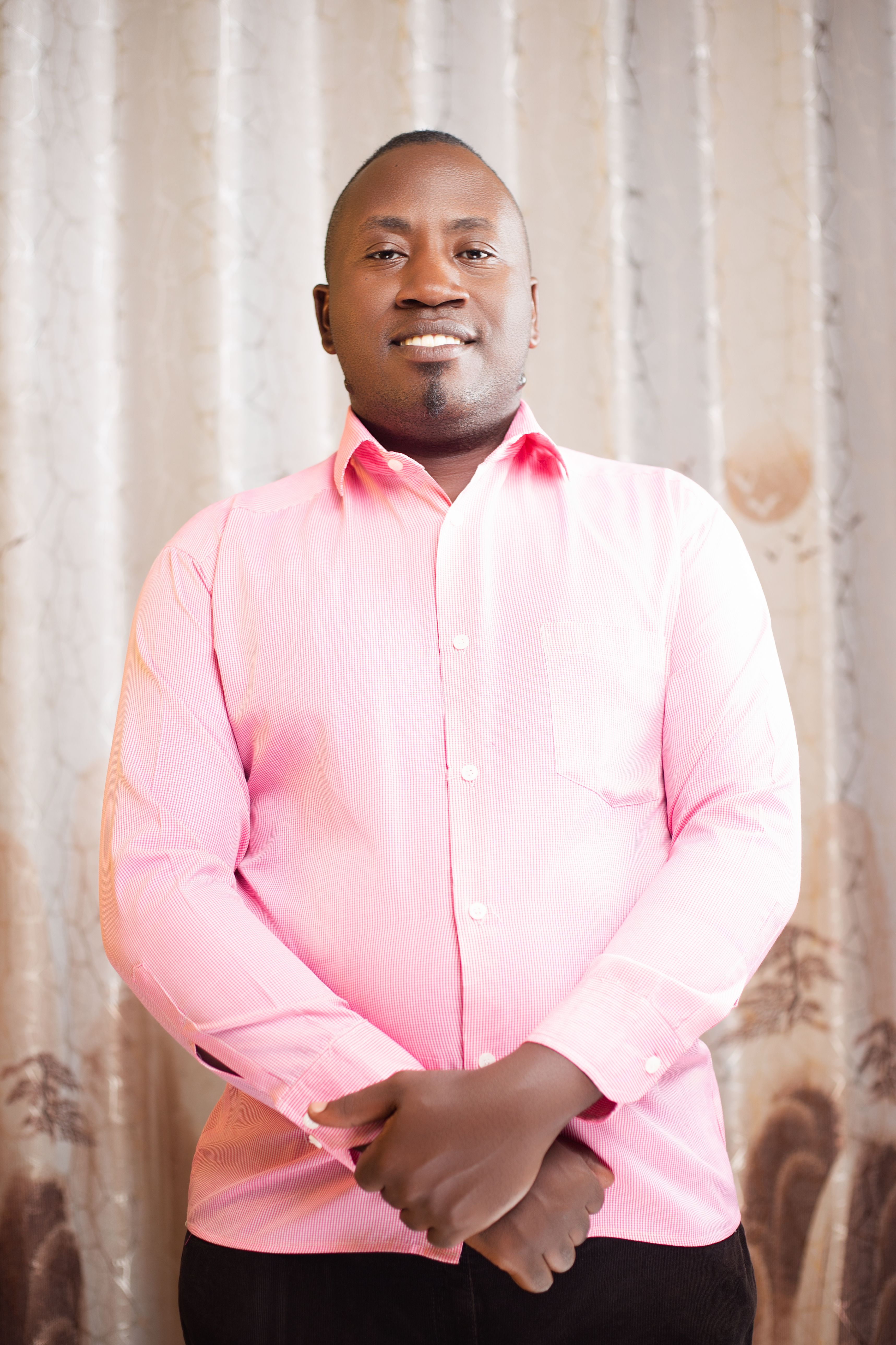 Portrait of a confident man wearing a pink shirt, standing with arms crossed indoors.
