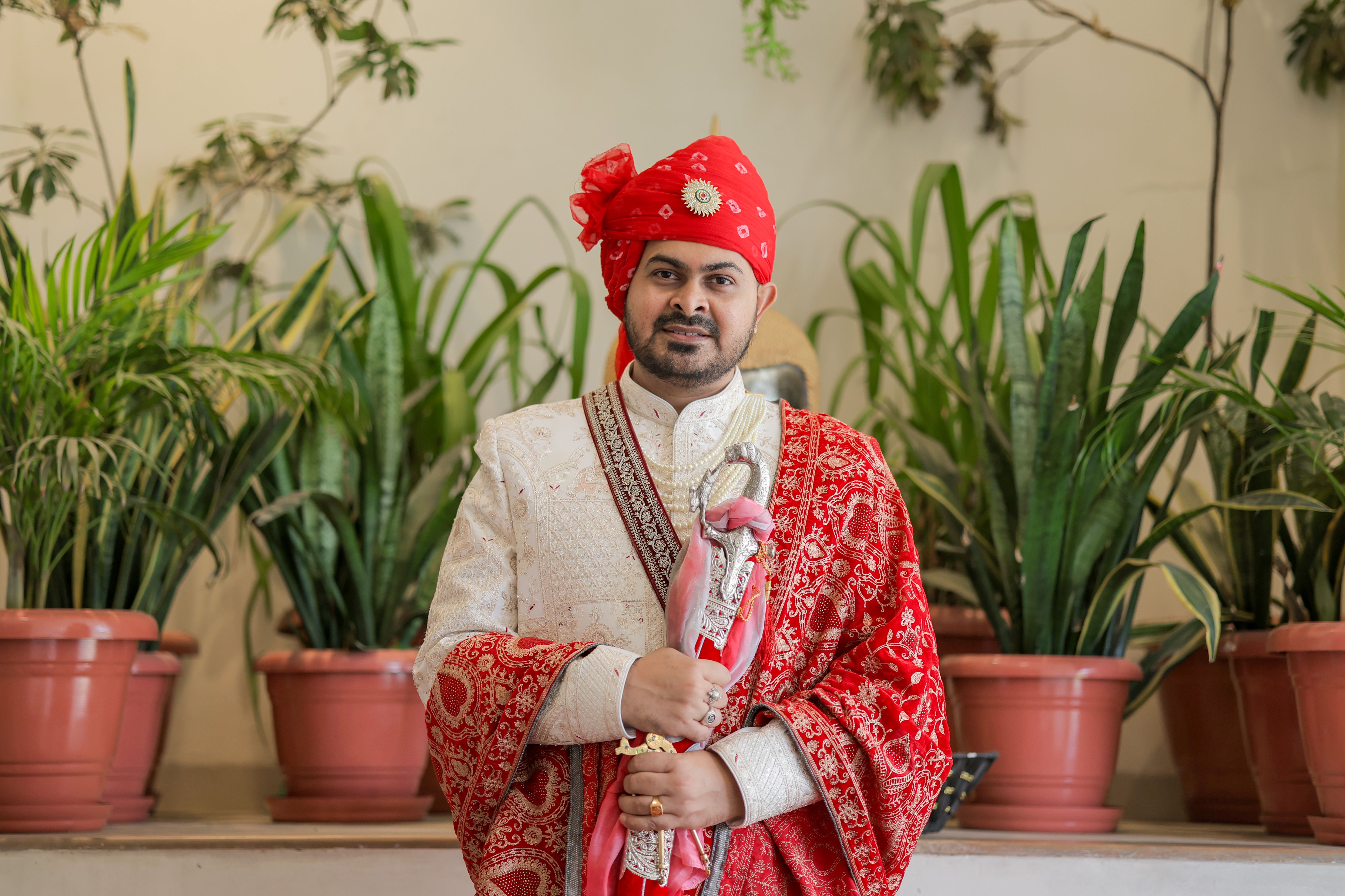 A stylish Indian groom dressed in traditional attire poses indoors with lush green plants.