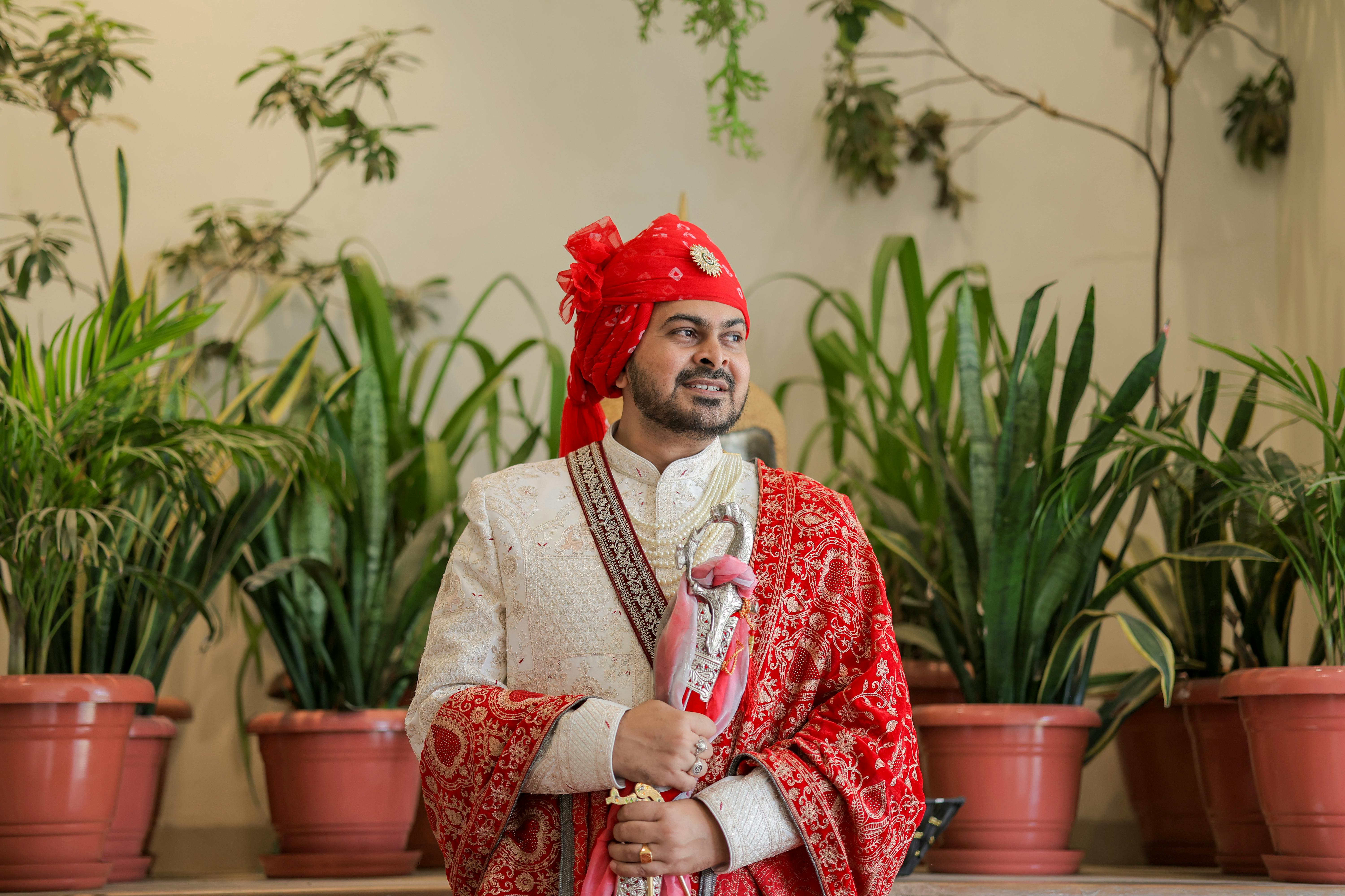 Indian groom in ornate attire with red turban posing indoors with greenery.