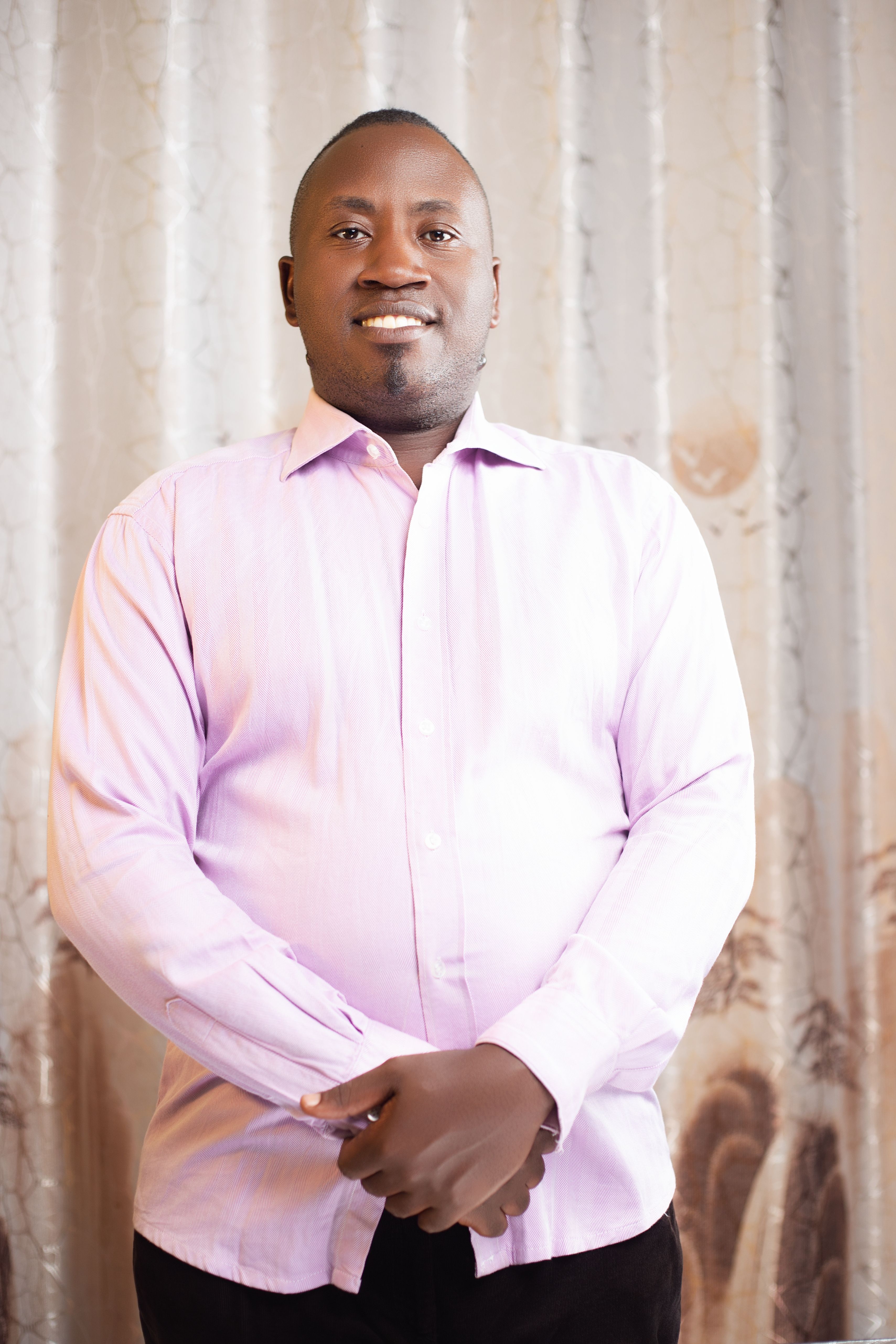 Portrait of a smiling man wearing a light pink shirt, standing in a well-lit indoor setting.