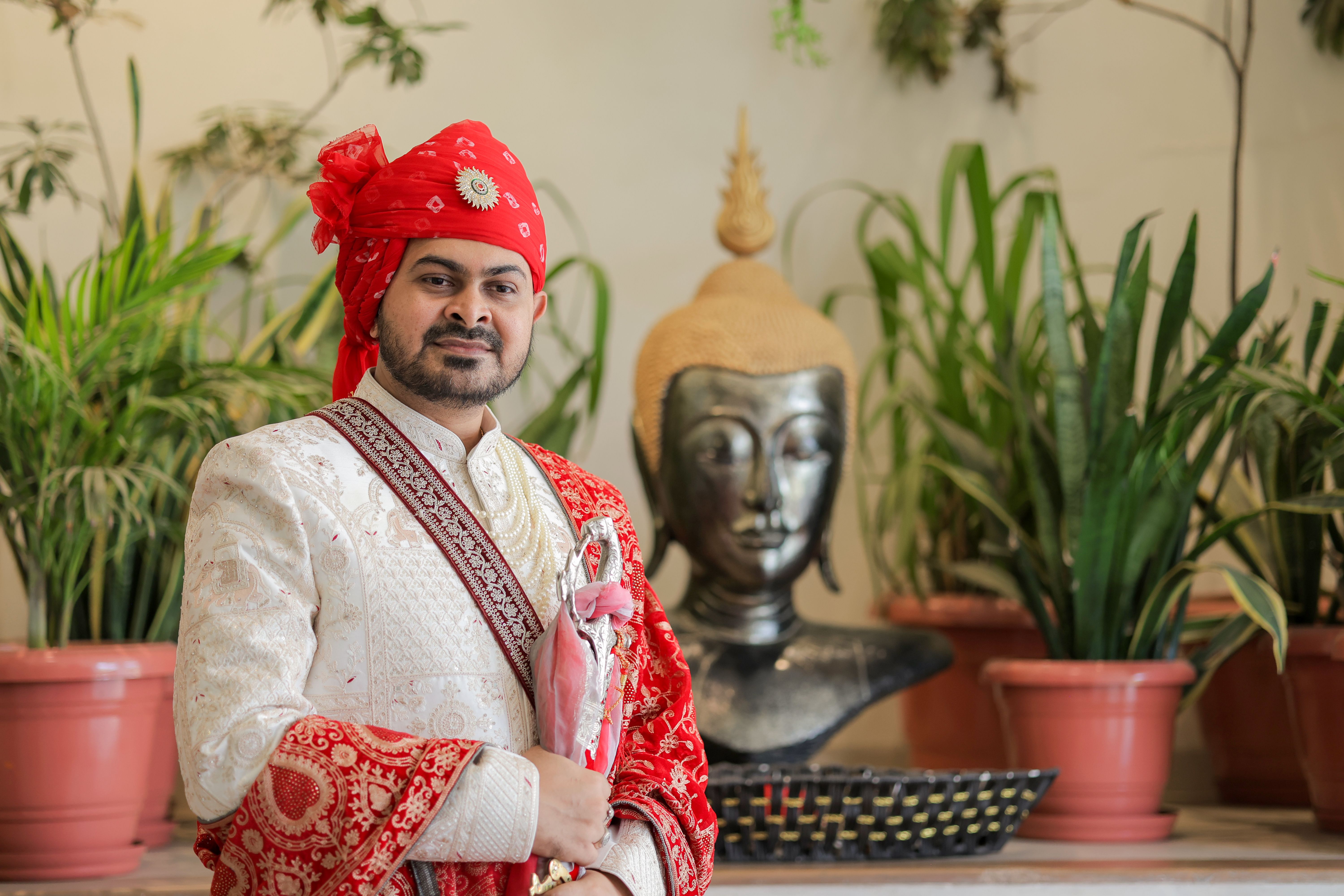 Indian groom in traditional attire with red turban and embroidered garments, standing by a Buddha statue.