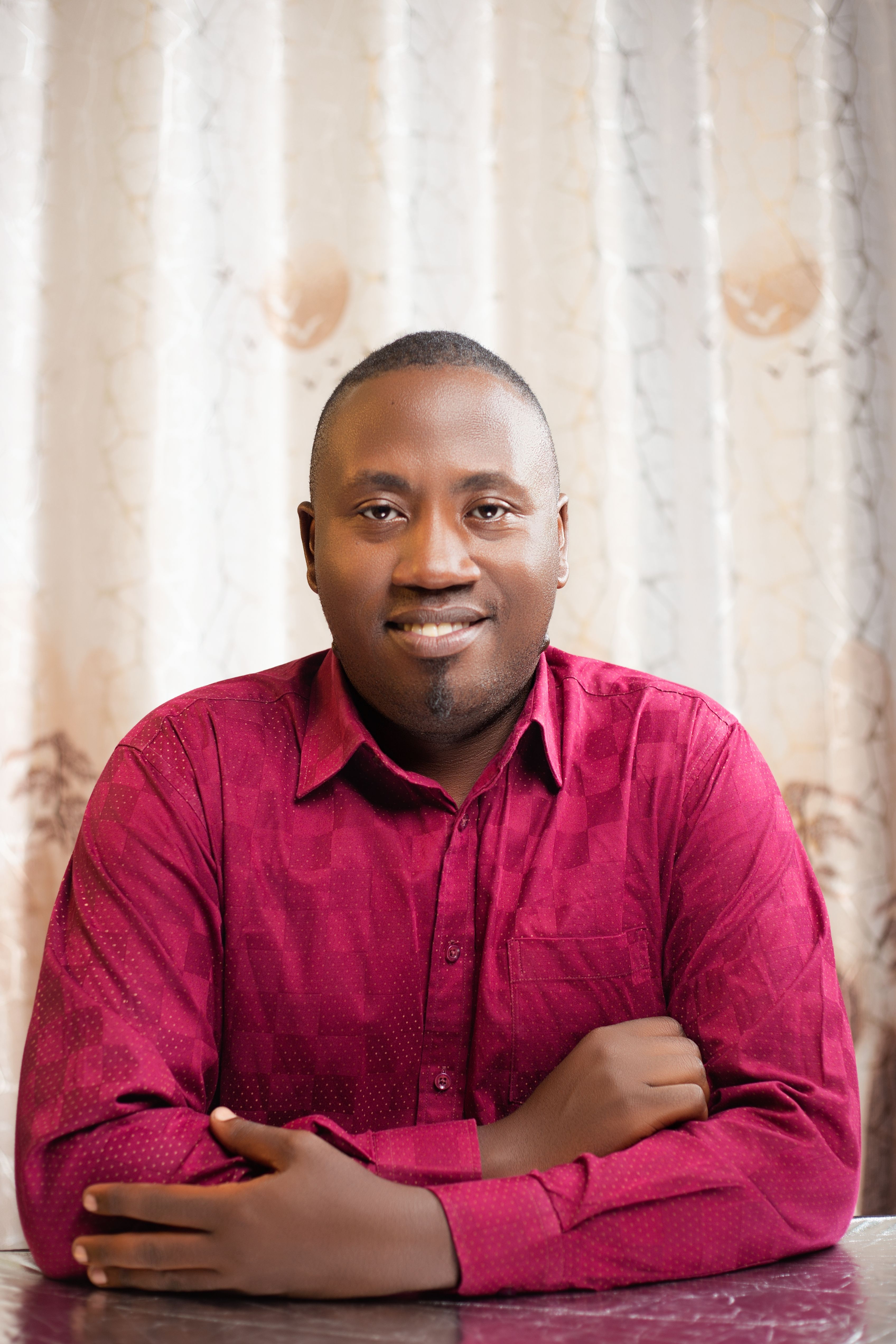 A cheerful man in a red shirt poses confidently indoors with a neutral curtain backdrop.