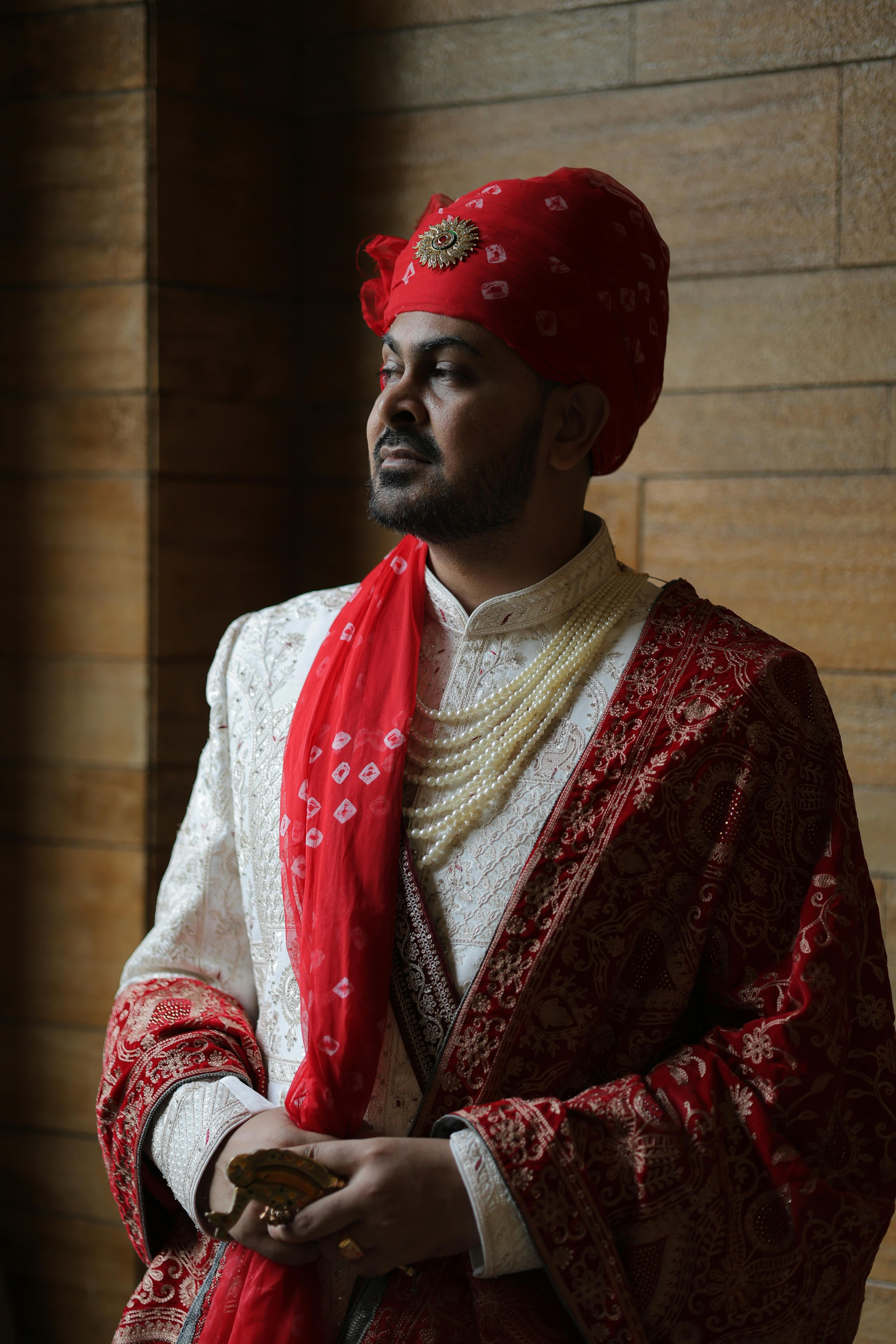 Indian groom in red and white attire, adorned with jewelry, posing indoors.