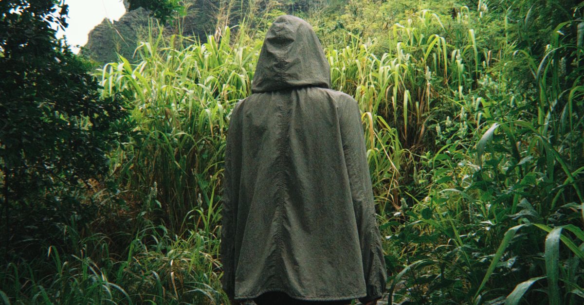 Photo by Caleb Lamb A lone hiker with a hood navigates a lush, overgrown jungle path in Hawaii, USA.