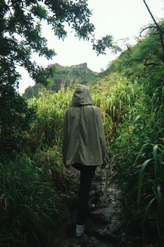 Photo by Caleb Lamb A lone hiker with a hood navigates a lush, overgrown jungle path in Hawaii, USA.
