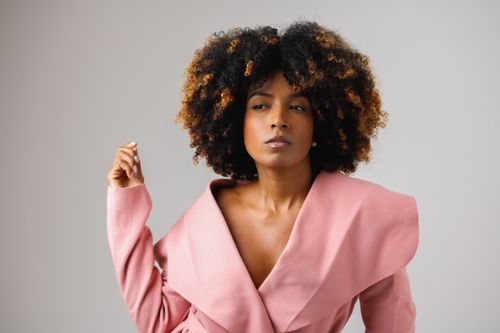 Free Stylish woman with curly hair posing confidently in a pink coat against a neutral background. Stock Photo