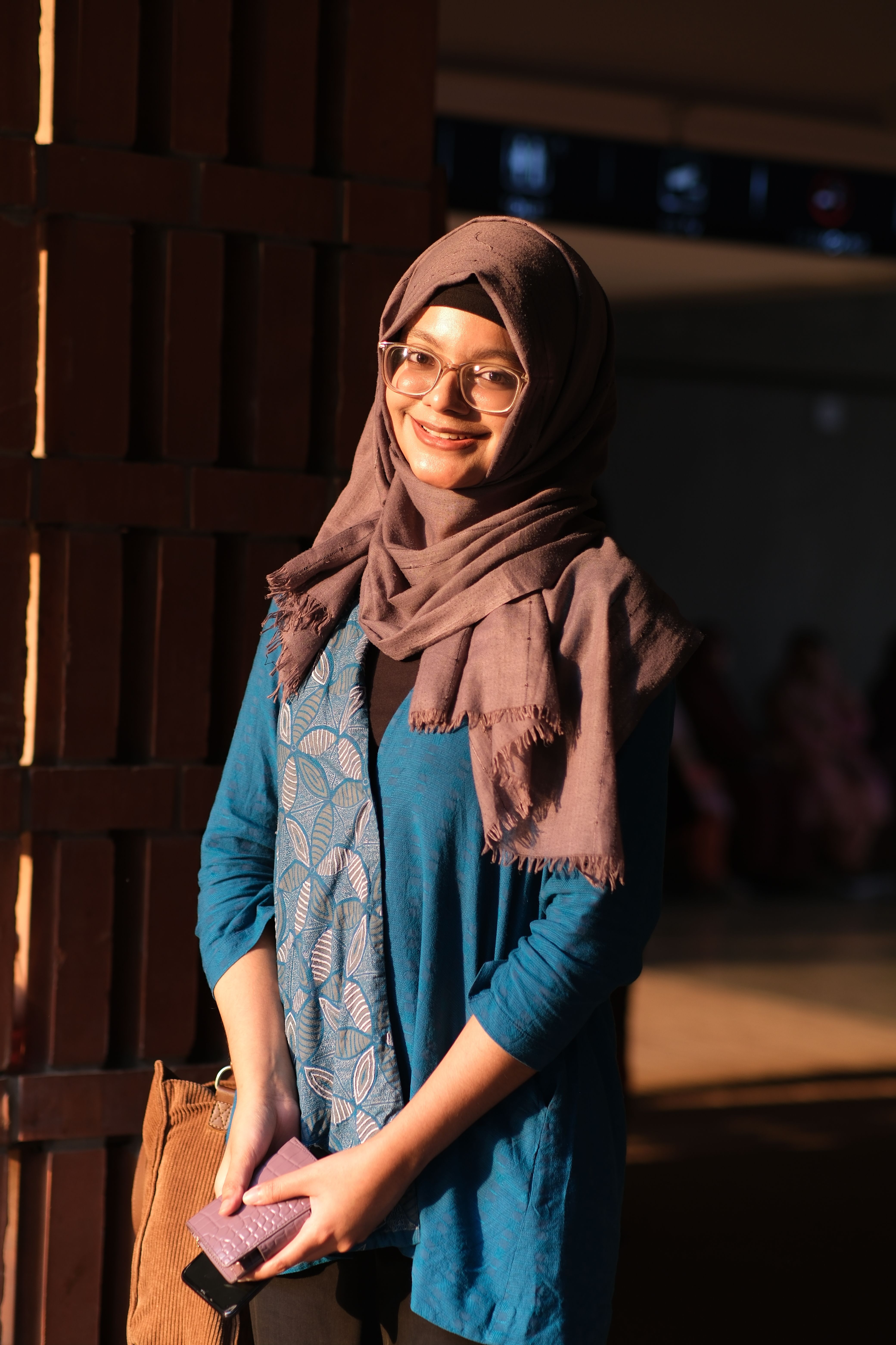 Portrait of a young woman smiling while wearing a hijab and glasses, captured in warm lighting outdoors.