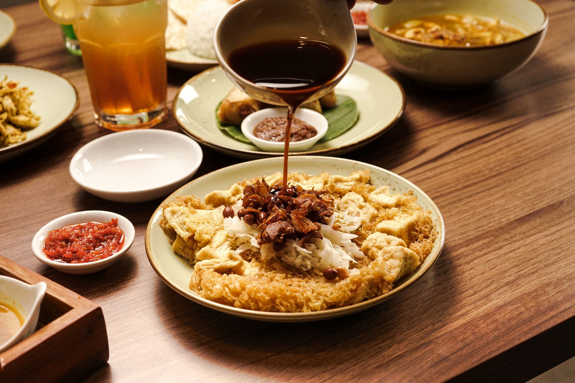 Indonesian dish with sauce being poured, served with spicy sambal on a wooden table.