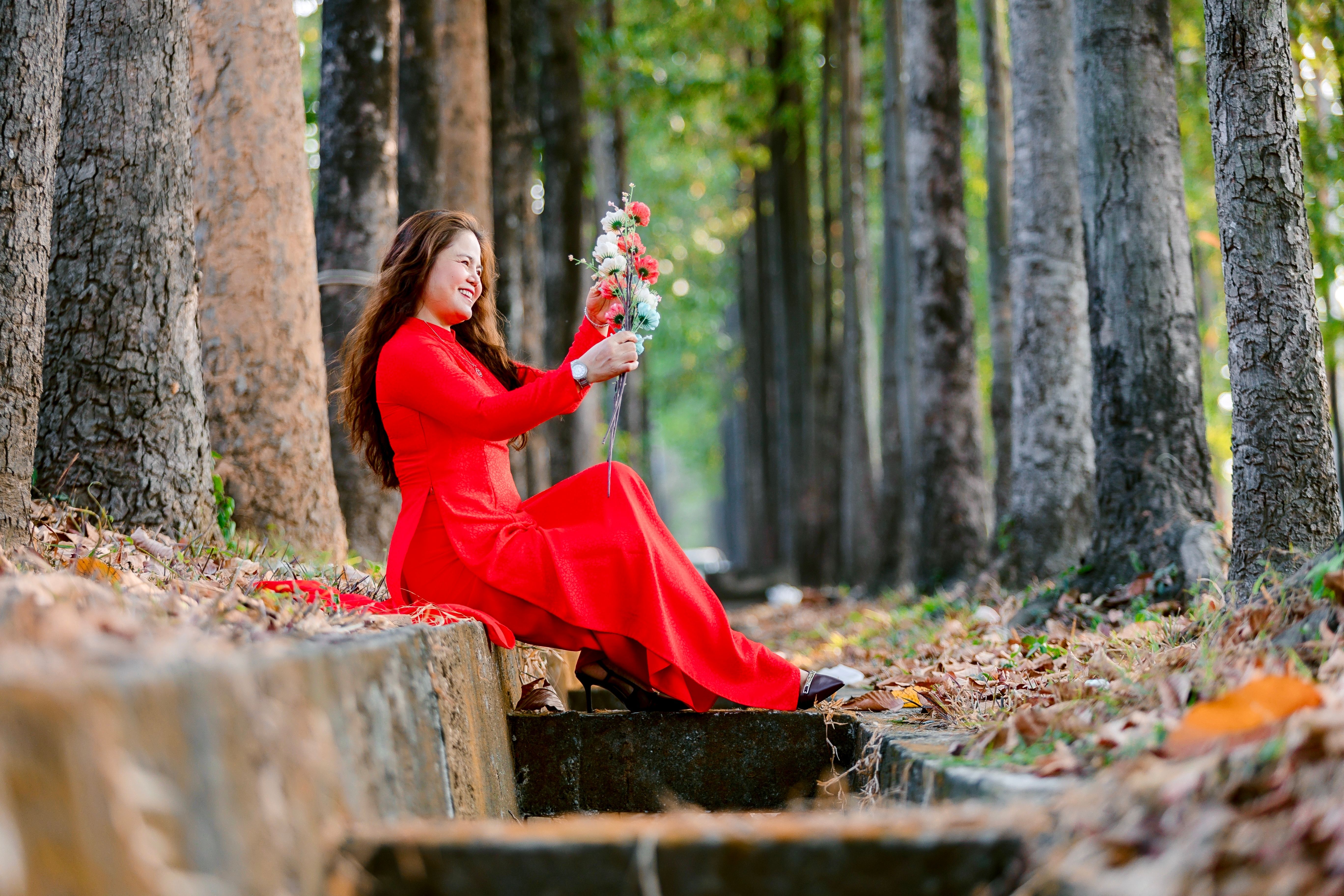 Smiling woman in red dress sitting with flowers in serene forest setting.