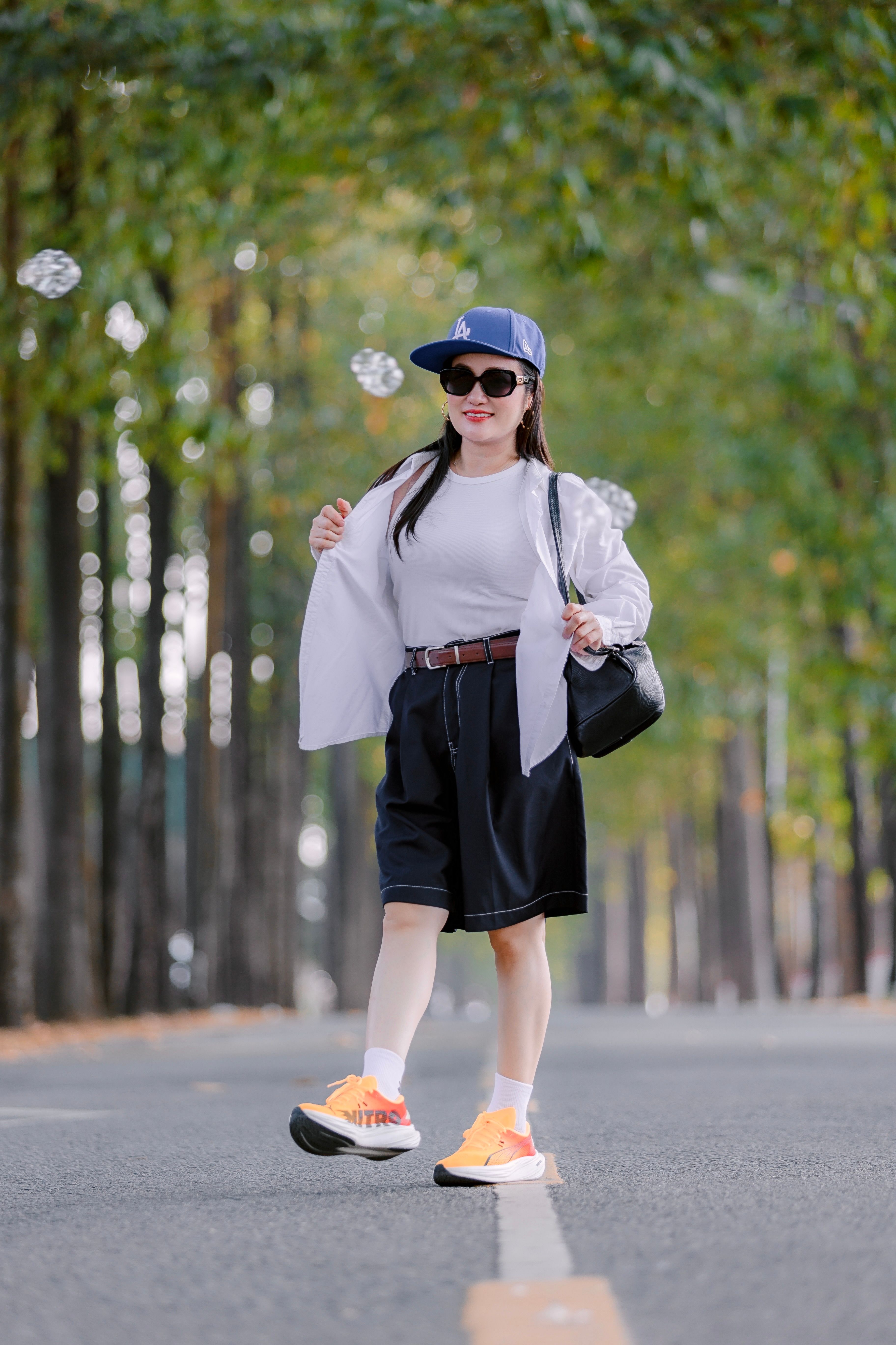 Stylish woman with sunglasses and cap walks confidently down an avenue lined with trees.