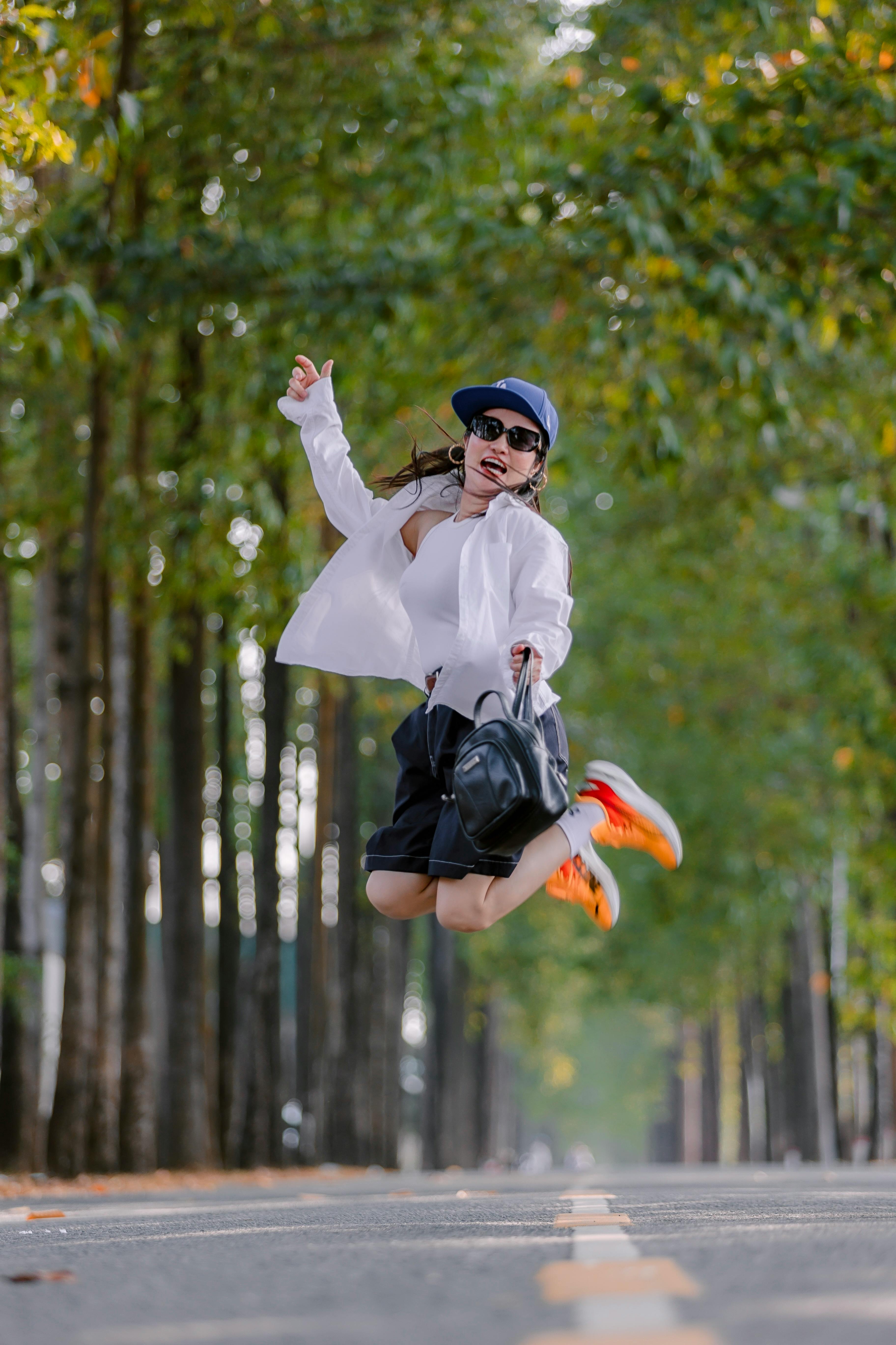 Woman in stylish outfit jumps energetically on a scenic, tree-lined street, embodying carefree joy.