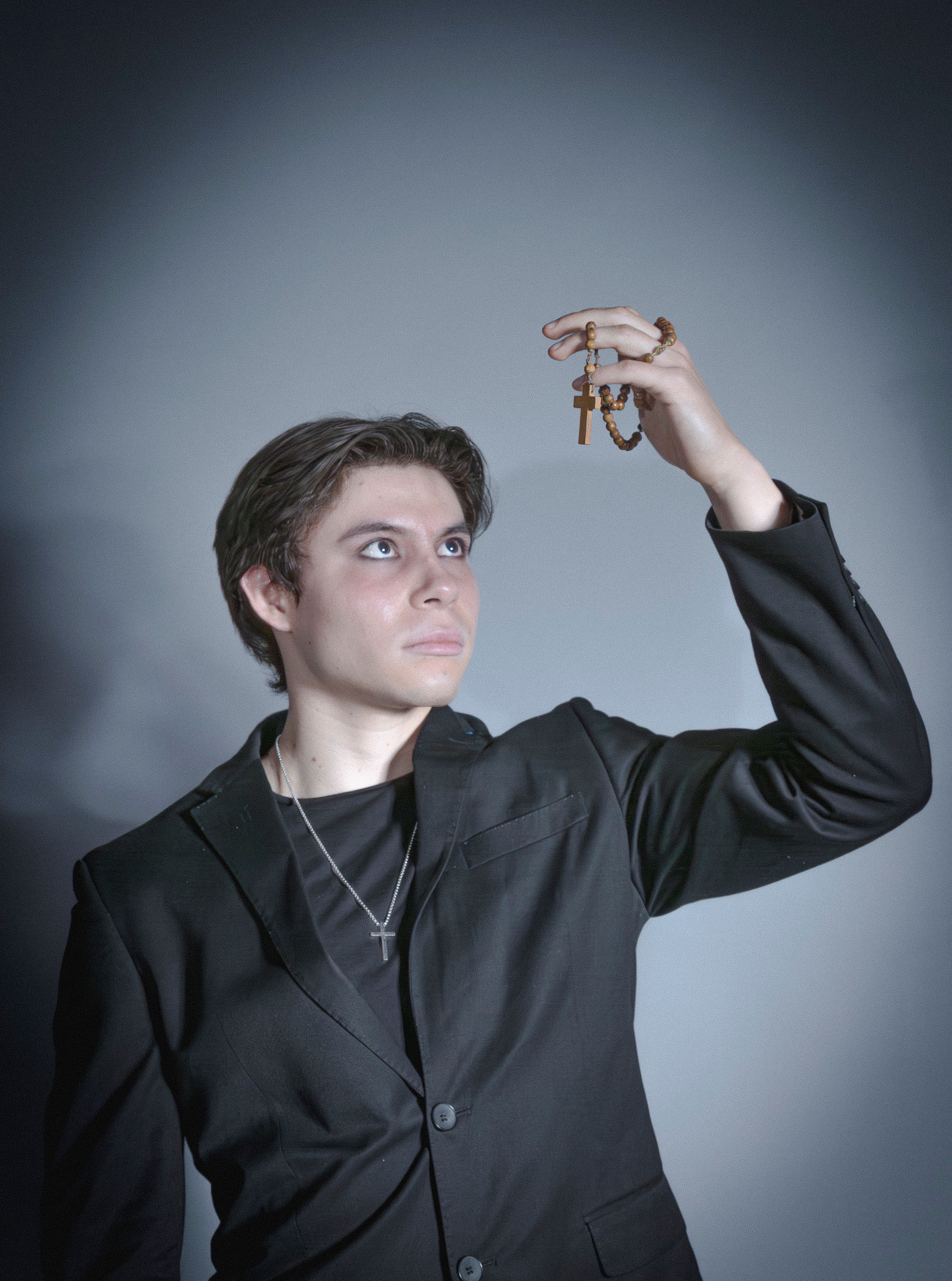 A man in a black suit holds a rosary with a crucifix. Dramatic lighting and expression.