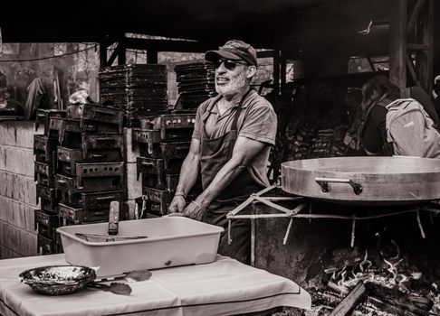 A smiling man cooks outdoors using traditional methods in a rustic setting.