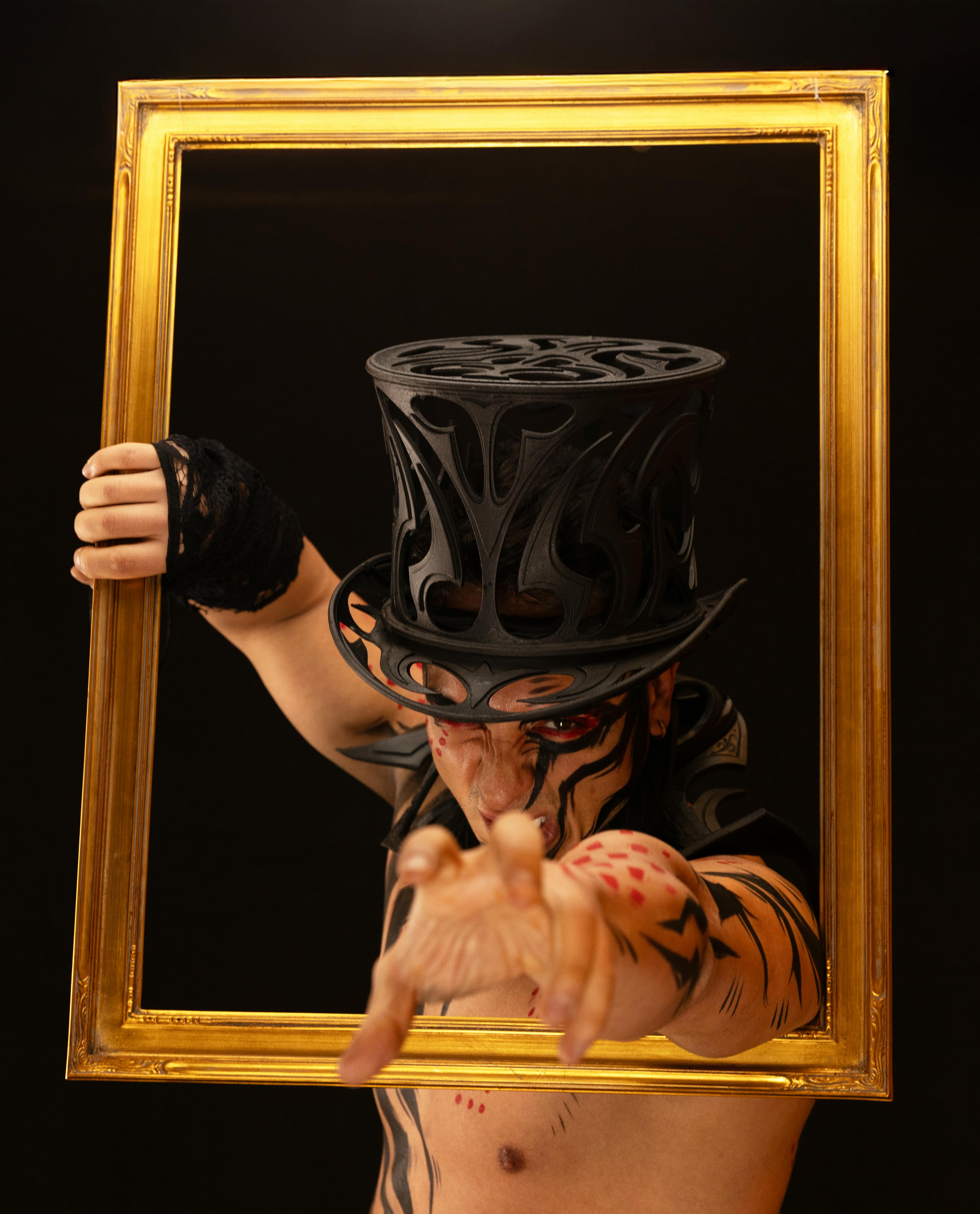 A person in artistic makeup poses with a frame and hat, against a dark background.