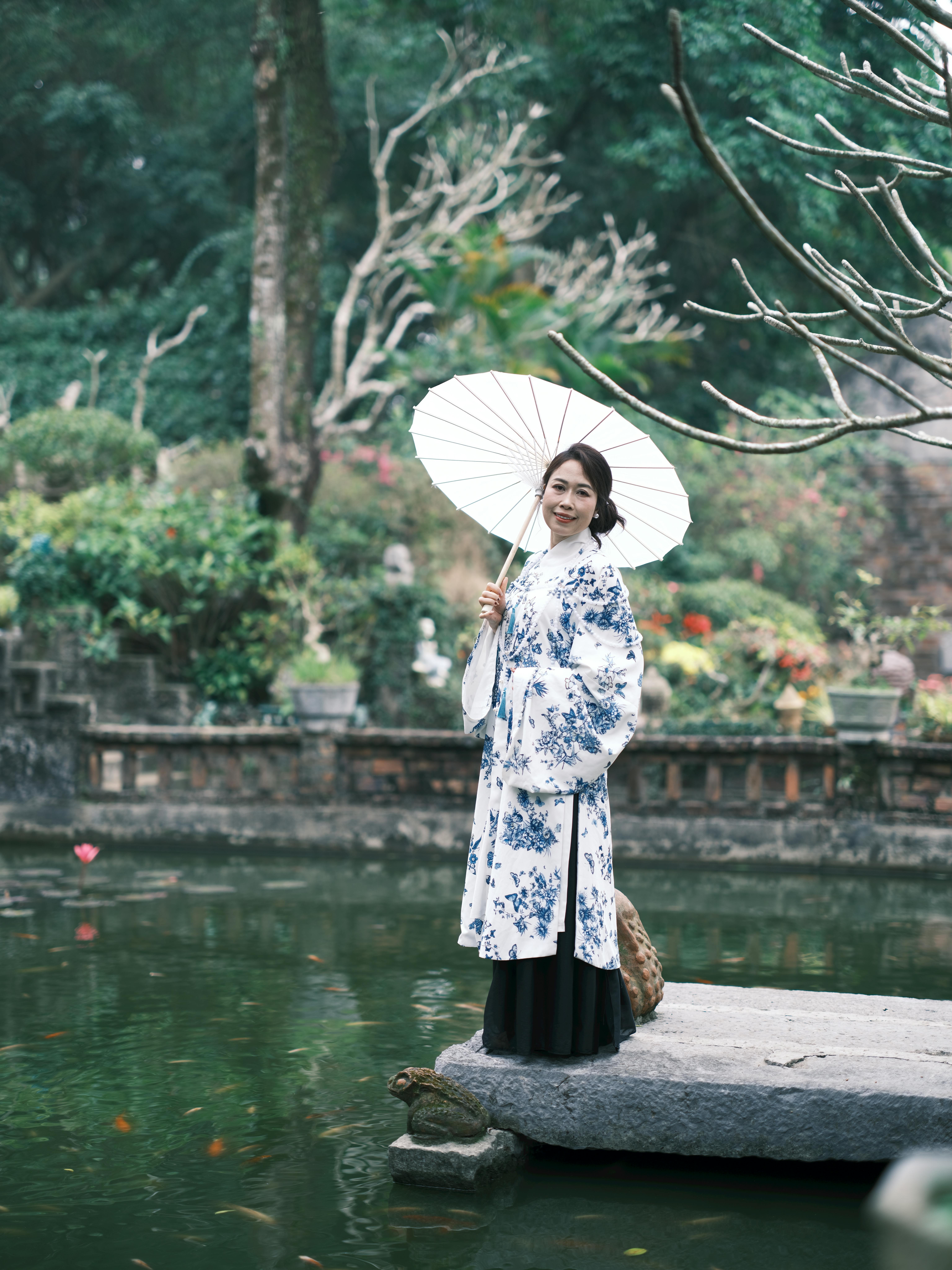 Elegant Vietnamese woman with parasol by a tranquil pond, showcasing traditional attire.