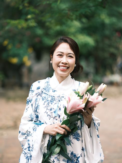 A cheerful woman in a floral Ao Dai holding lilies, outdoors in Việt Nam.
