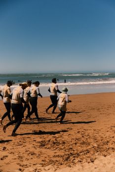 A group of people in wetsuits running along a sunny beach towards the ocean waves.