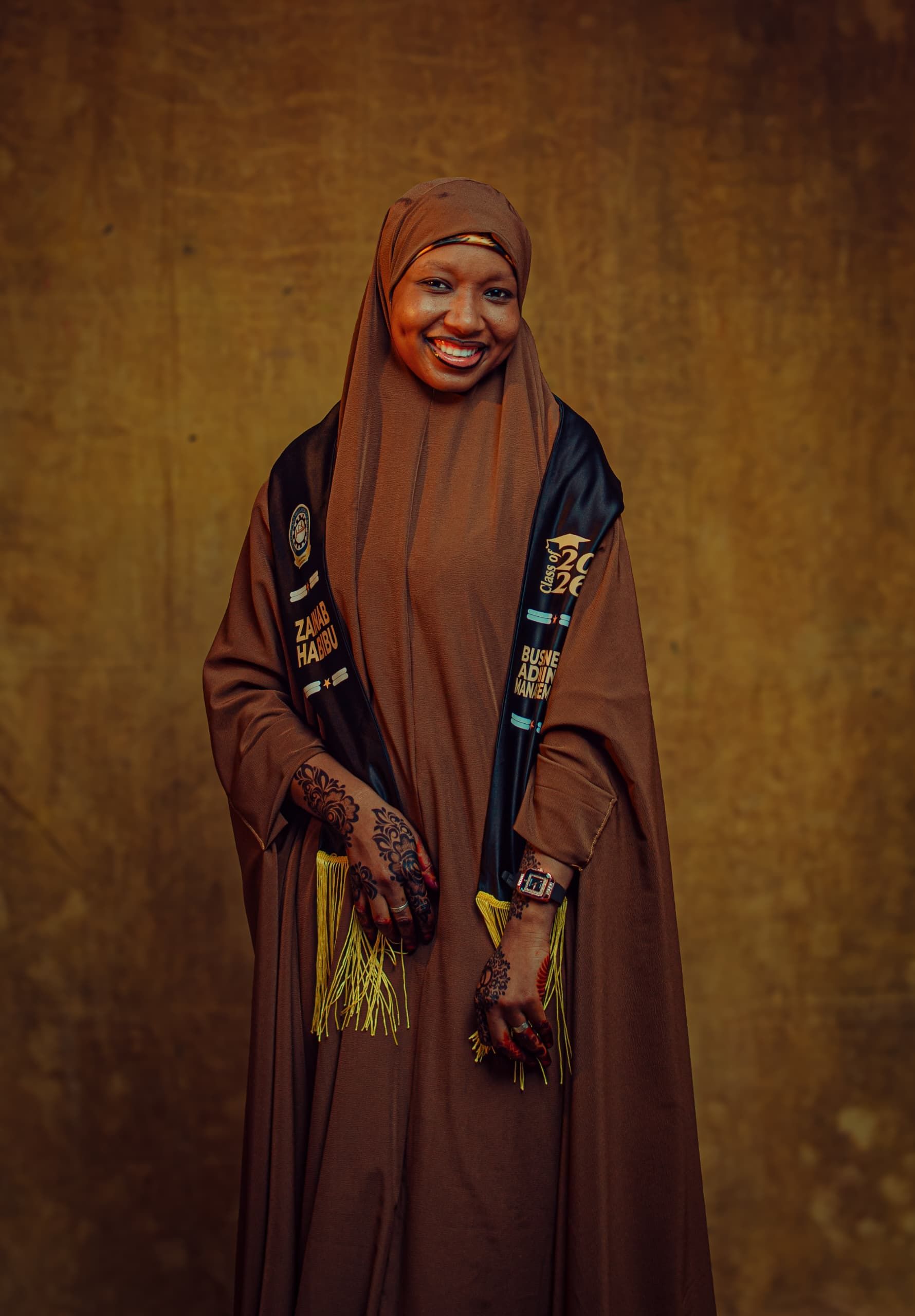 Smiling woman in brown traditional attire with henna and sash, studio portrait.