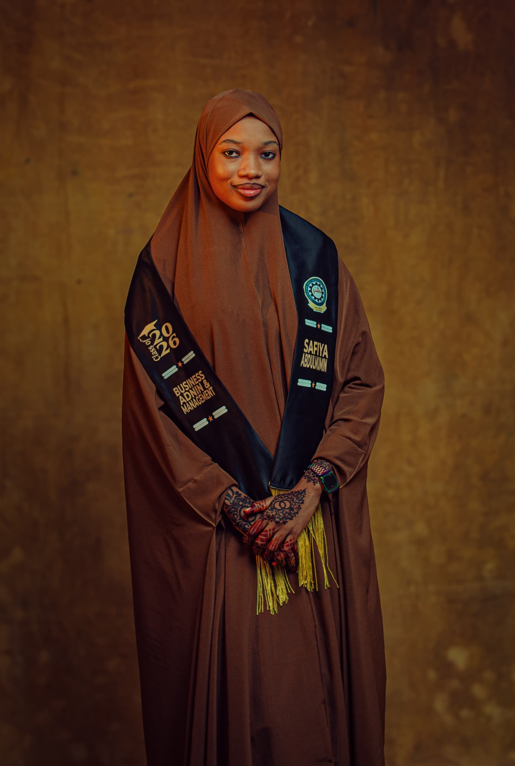 A woman in a hijab and graduation gown stands confidently with intricate henna on her hands.