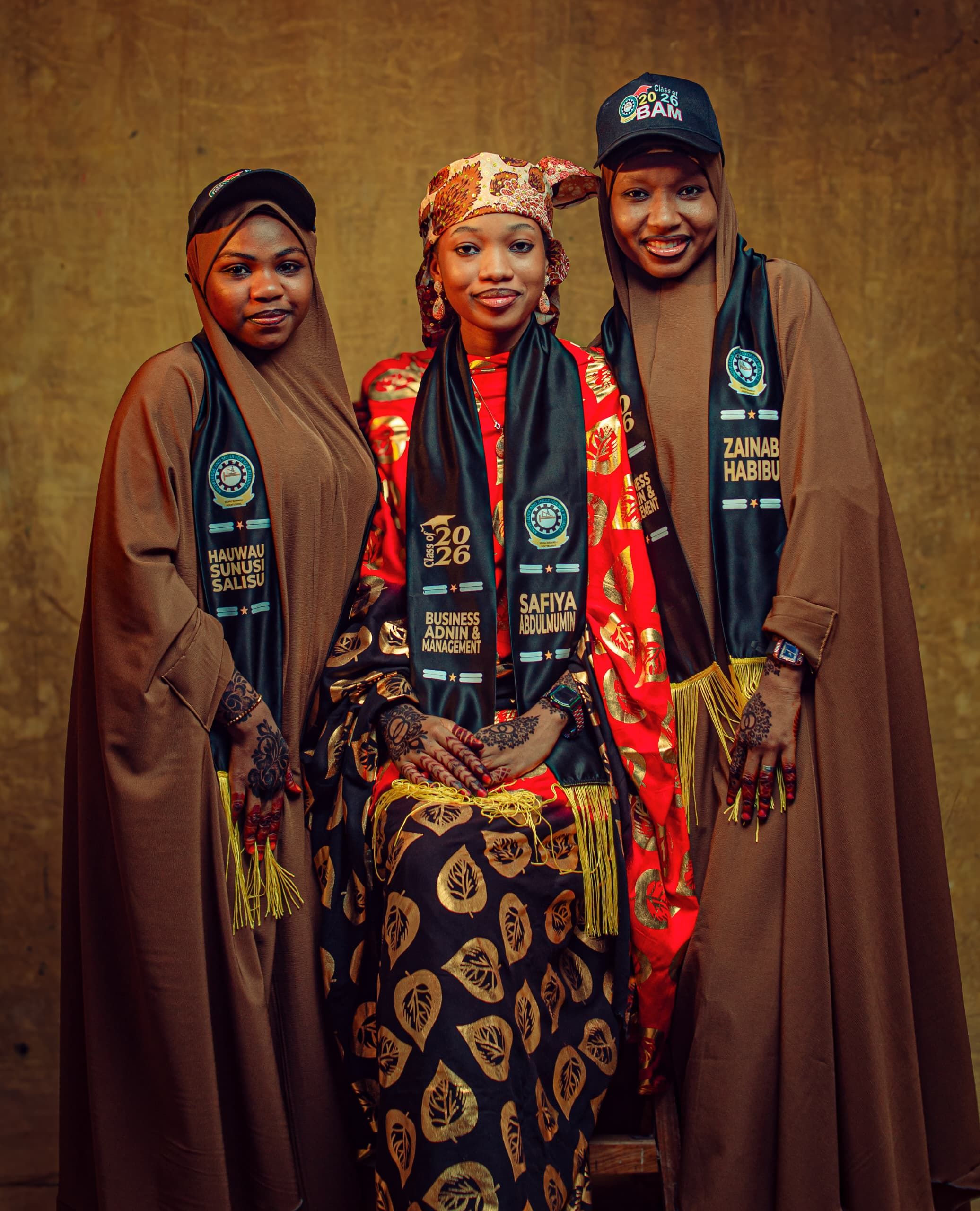 Three women posed in traditional clothing, celebrating graduation with black sashes.