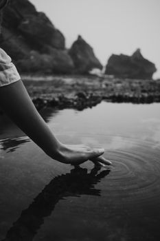 A black and white photo of a hand gently touching water near rocks, creating ripples.