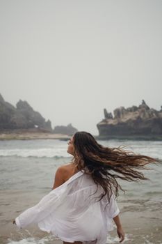 A young woman in a white dress enjoys the ocean waves on a misty beach.