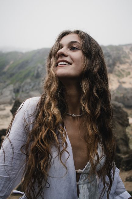 Smiling woman with long wavy hair in a white shirt, enjoying the outdoors. Scenic rocky landscape in the background.