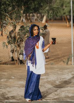 A fashionable young woman in colorful Nigerian attire holds a cup outdoors.