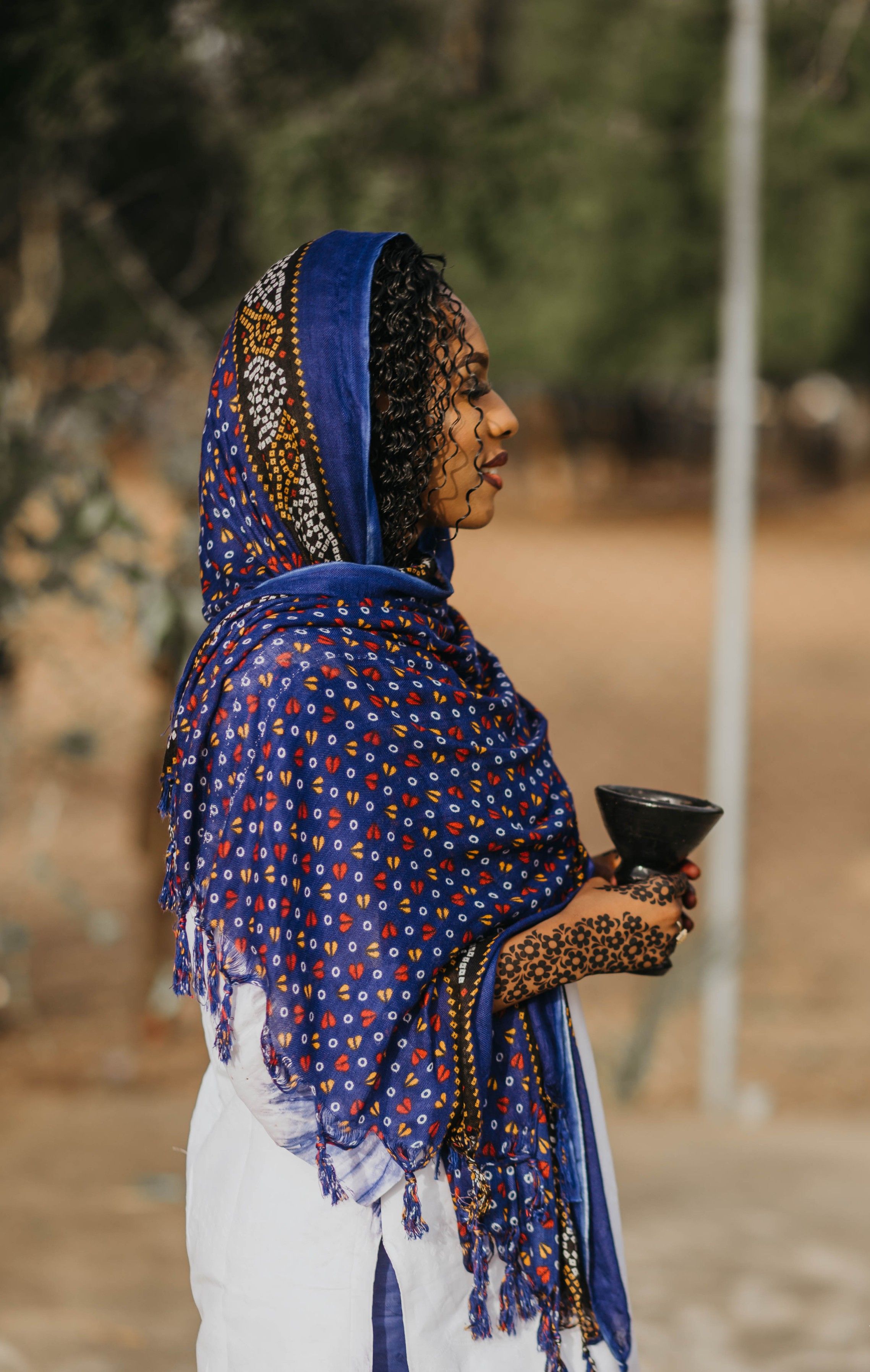 Profile of an African woman holding a cup outdoors, wearing colorful traditional attire.