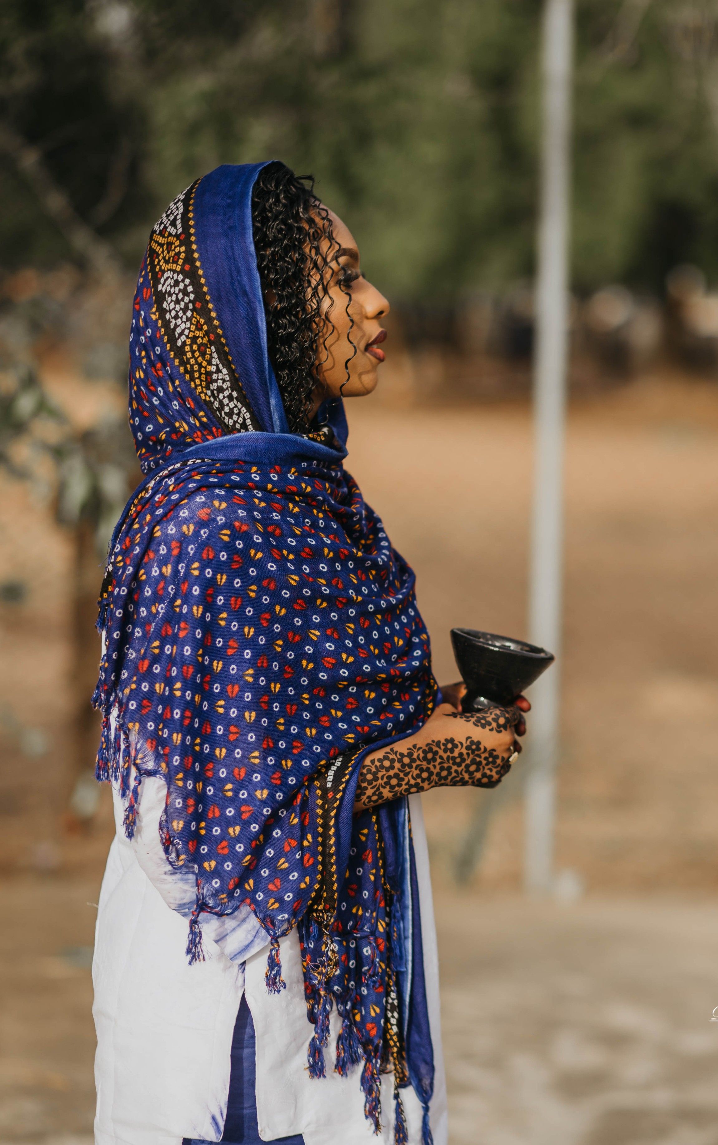 Side view of a Nigerian woman in colorful traditional clothing, holding a pot outdoors.