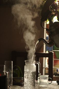 A kettle pouring steaming hot water into a glass jug on a kitchen table, indoors.